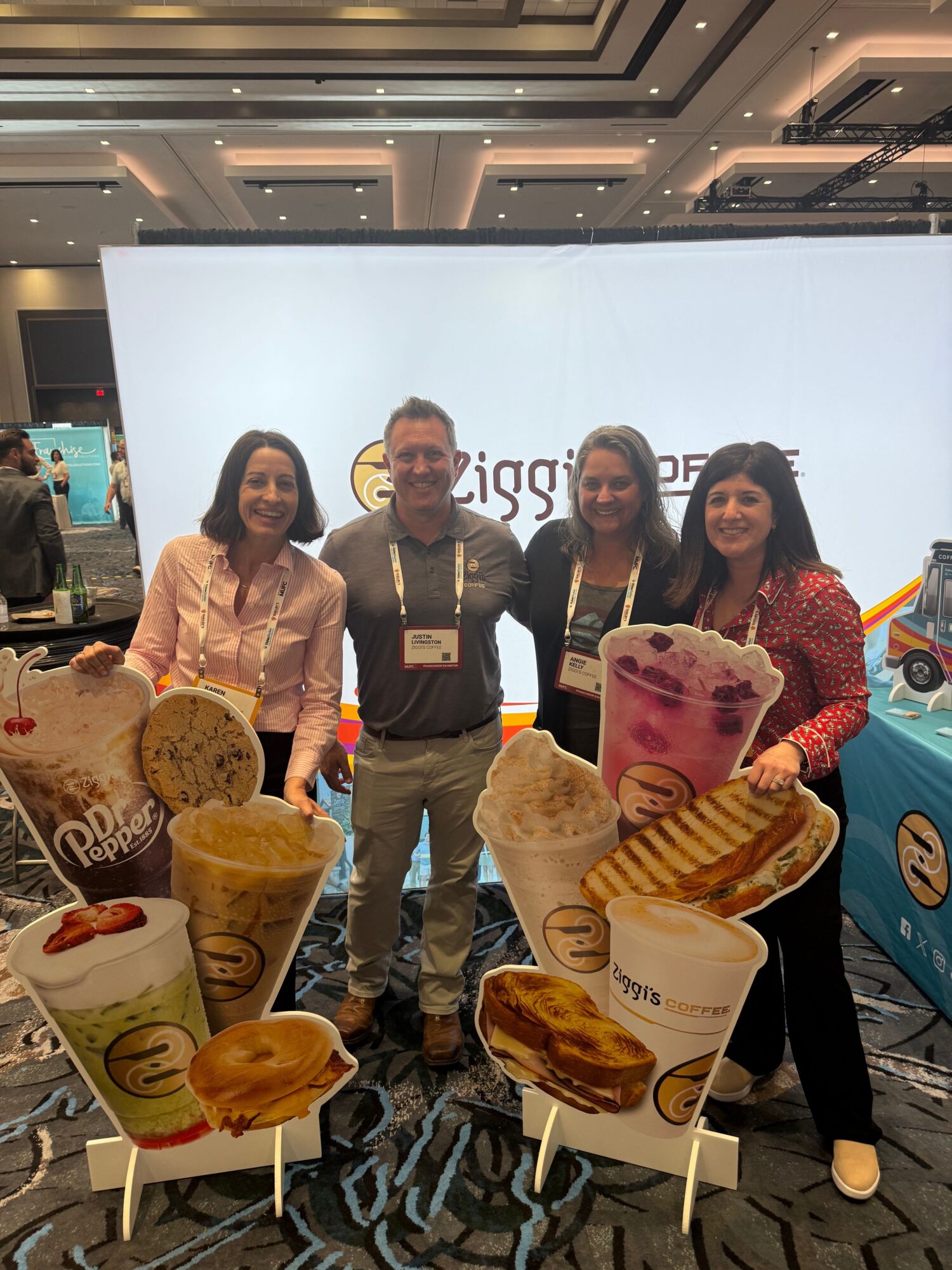 Four people standing in front of a large screen with ice cream cones and cups displayed, inside a shopping mall or store.