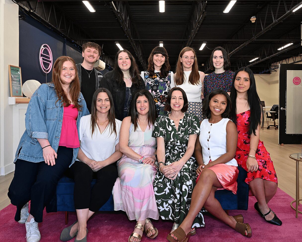 Group of ten women and one man sitting and standing in a room with black ceiling, pink carpet, and a blue sofa.