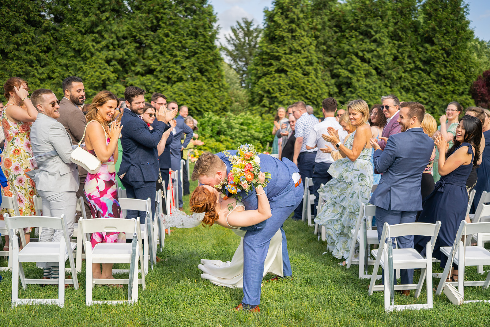 People standing outdoors in a garden, clapping and taking photos during a wedding ceremony, with trees in the background.