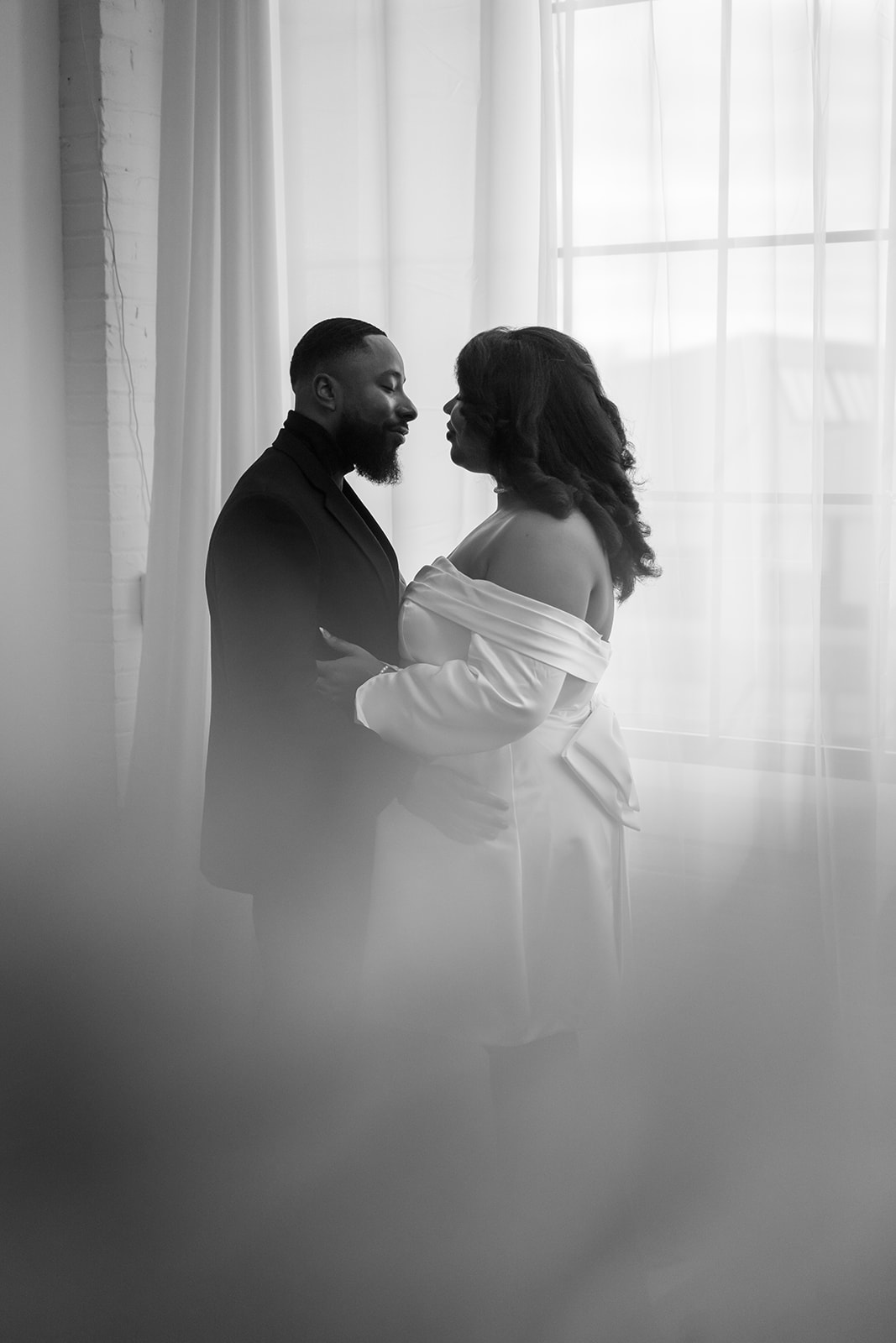 Two people face each other near a window with curtains, holding hands, in black and white.