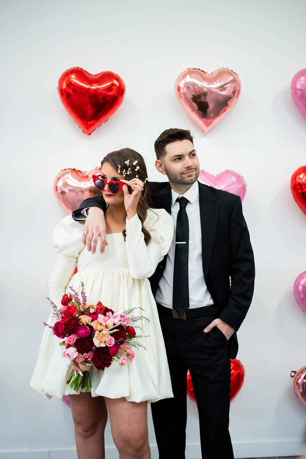 A woman in a white dress holding a bouquet of pink and red flowers, with a man in a black suit standing beside her, against a backdrop of heart-shaped balloons in red, pink, and purple.