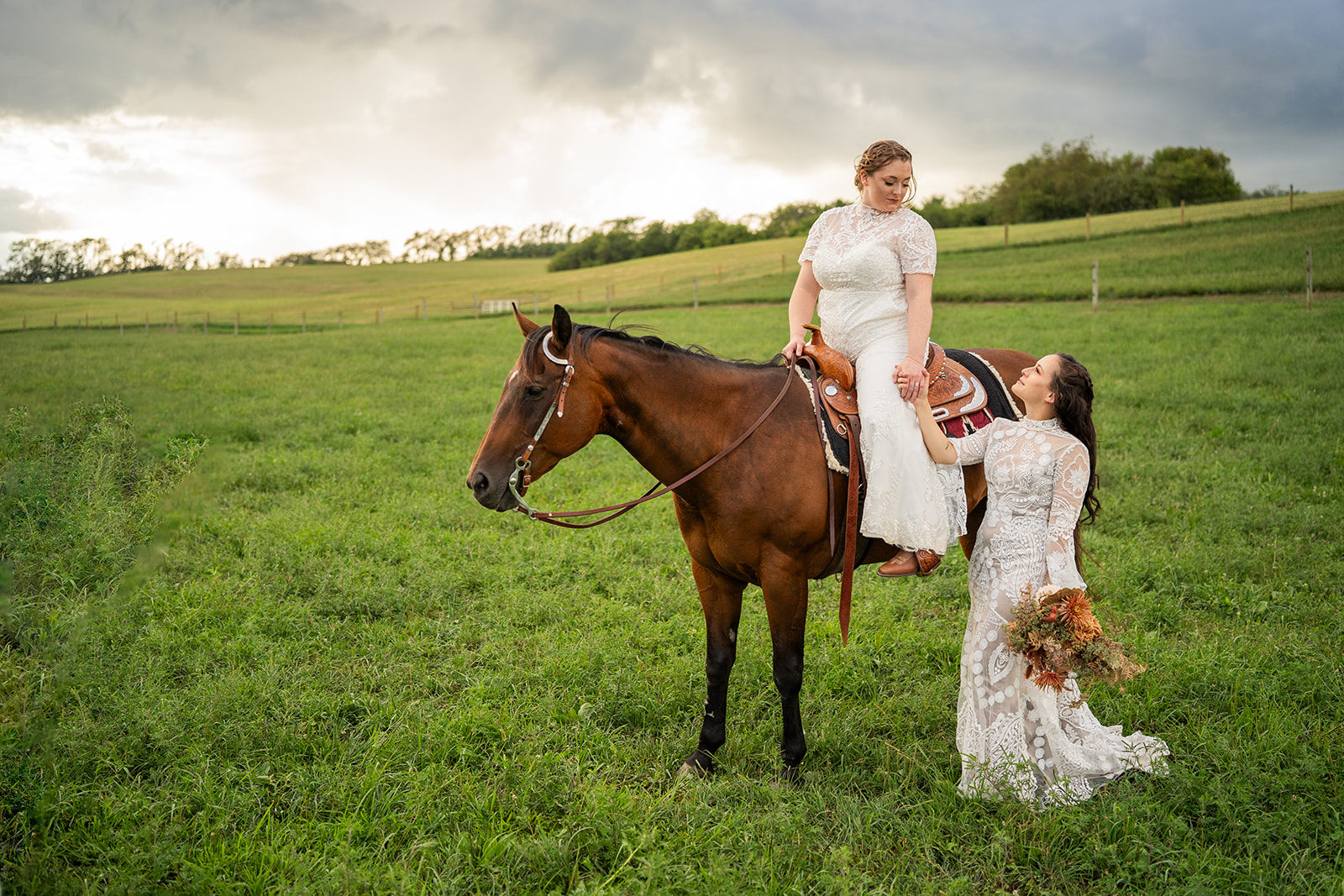 Two women in white dresses with flowers, one on a horse and one standing, in a green field under cloudy sky
