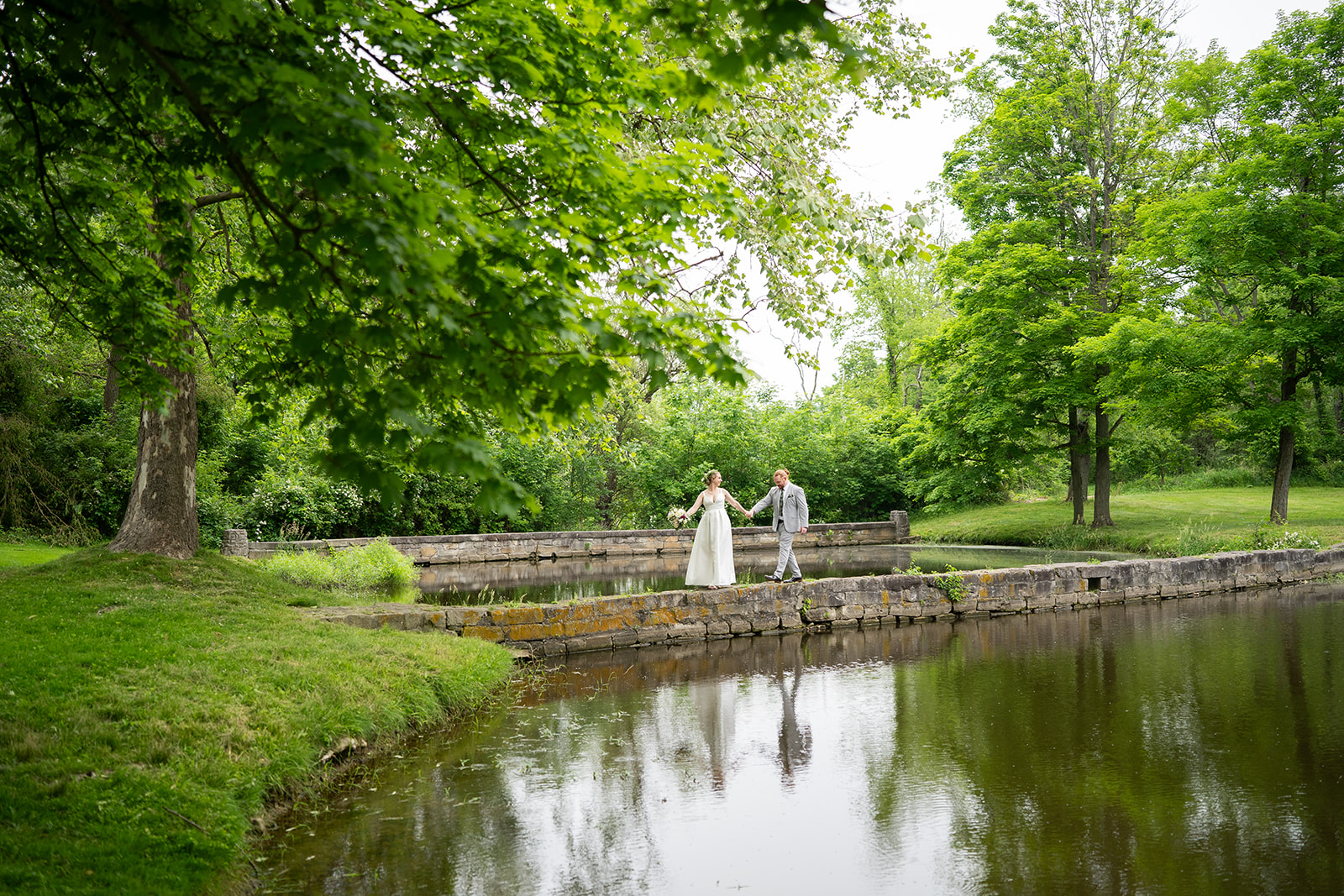 Two people stand on a stone ledge near a pond in a lush green park, holding hands and facing each other.