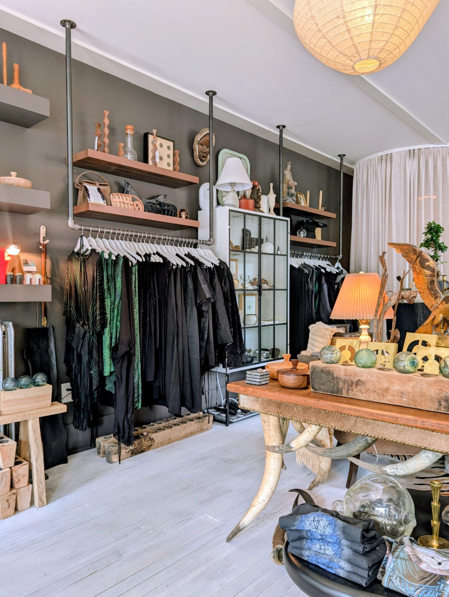 Clothing rack with black garments, shelves with decorative items, glass cabinet, and a wooden table with teapots and a lamp.