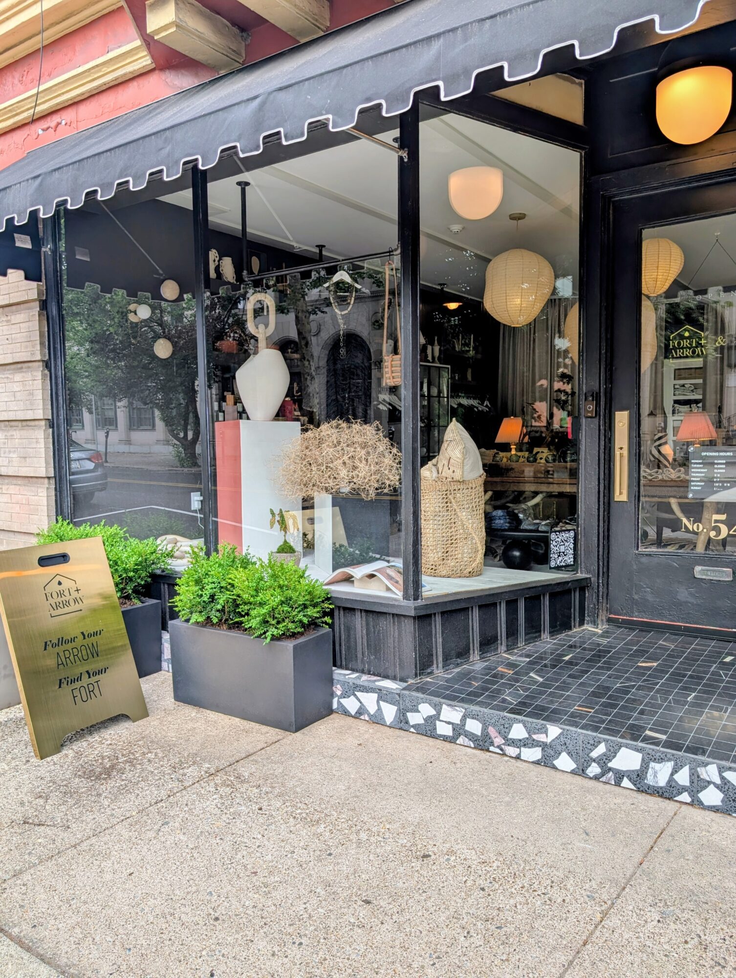 Storefront with large glass windows displaying decorative vases and plants, black awning, and sidewalk outside.