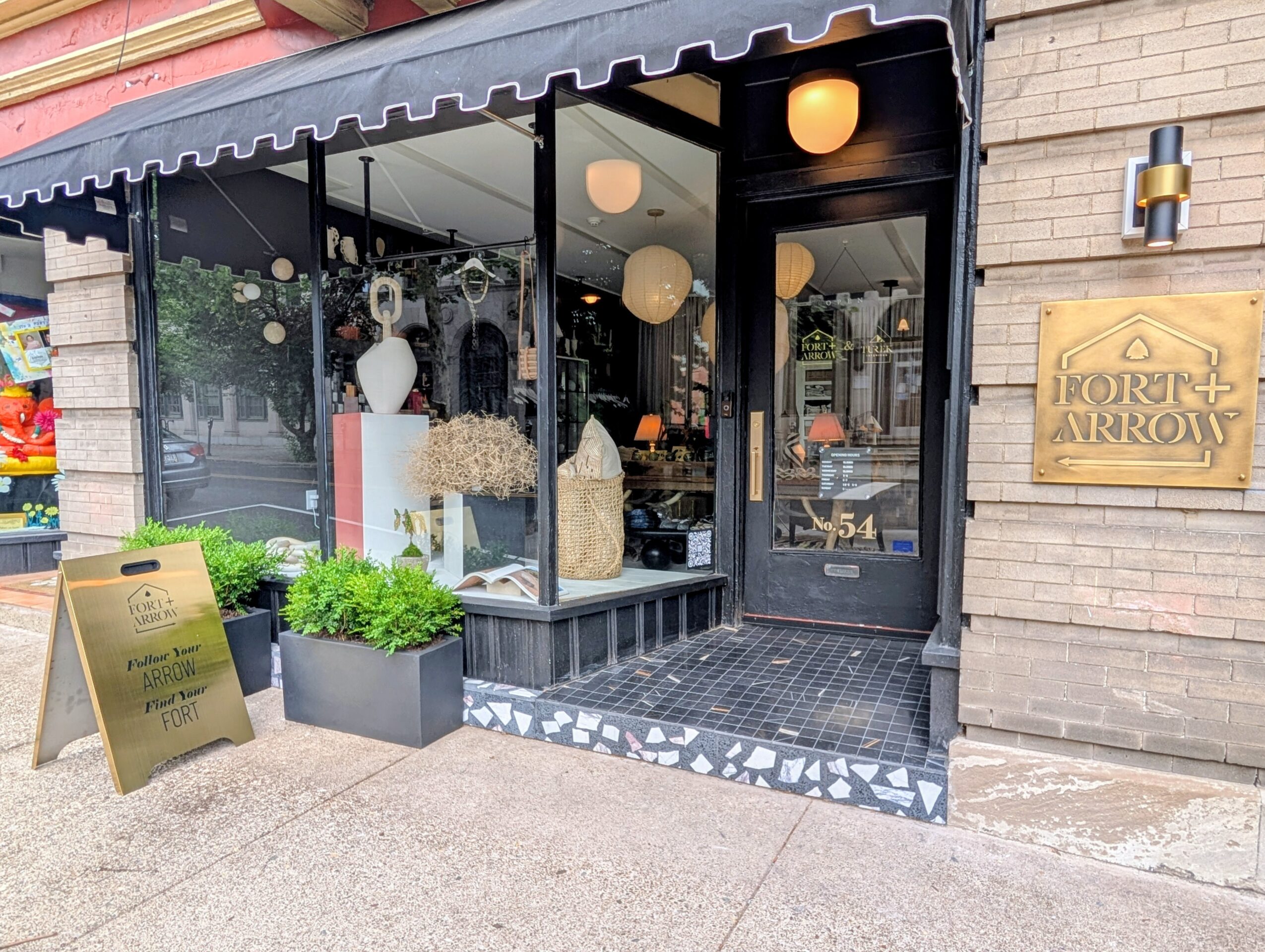 Storefront with glass windows, potted plants outside, and a sign on the brick wall, with interior lighting visible.