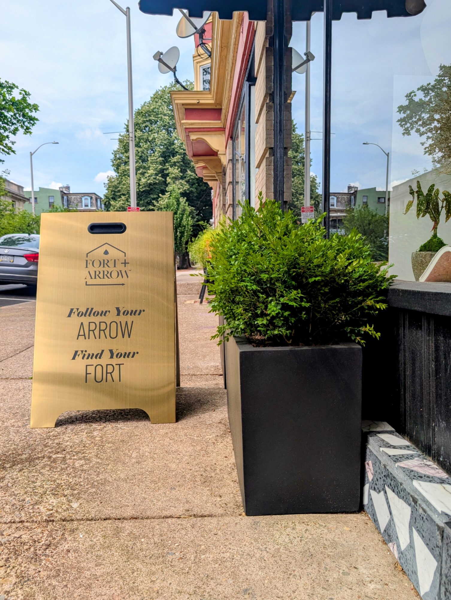 Sidewalk with a signboard and a large potted plant outside a building, with trees and a blue sky in the background.