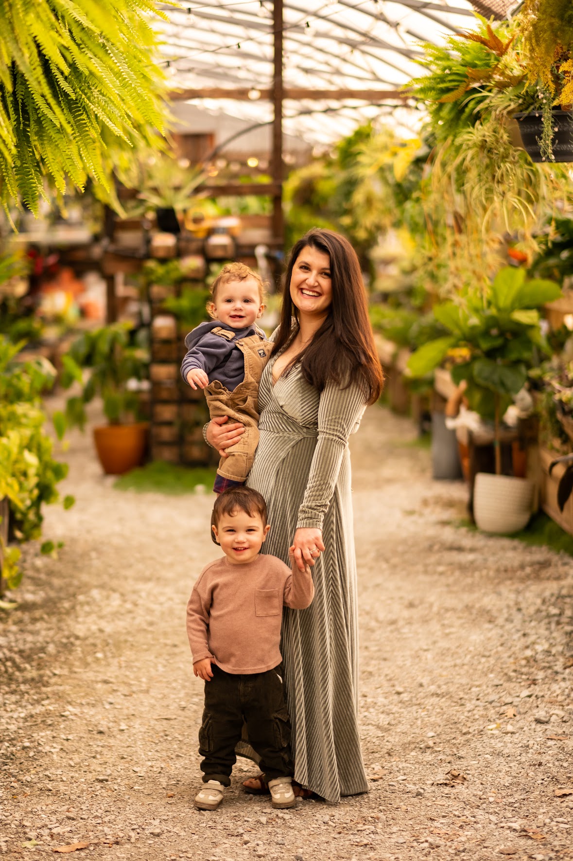 Woman holding a baby in a greenhouse with plants and hanging greenery, smiling, with a young boy standing nearby, all smiling.