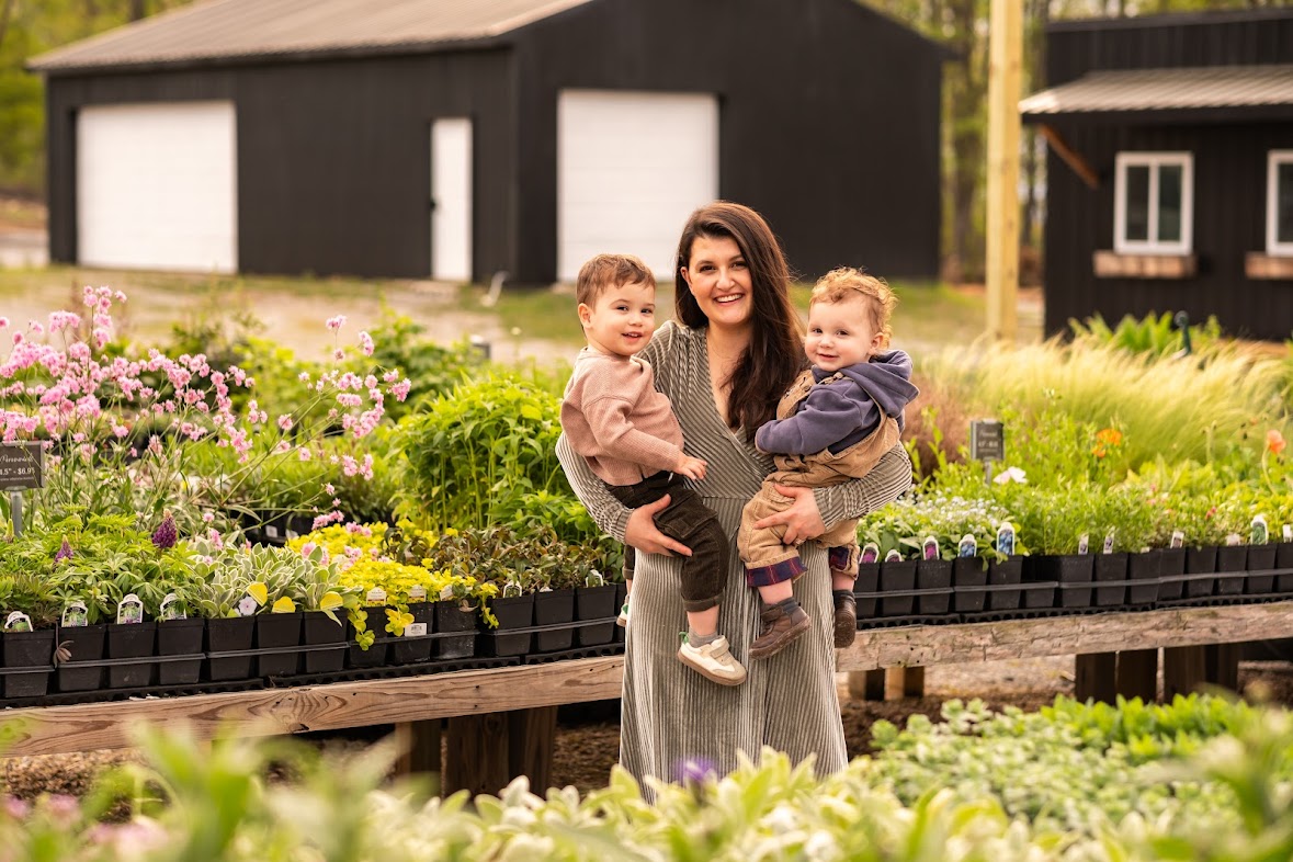 Woman holding two children in a garden with colorful flowers and greenery, black buildings in the background.