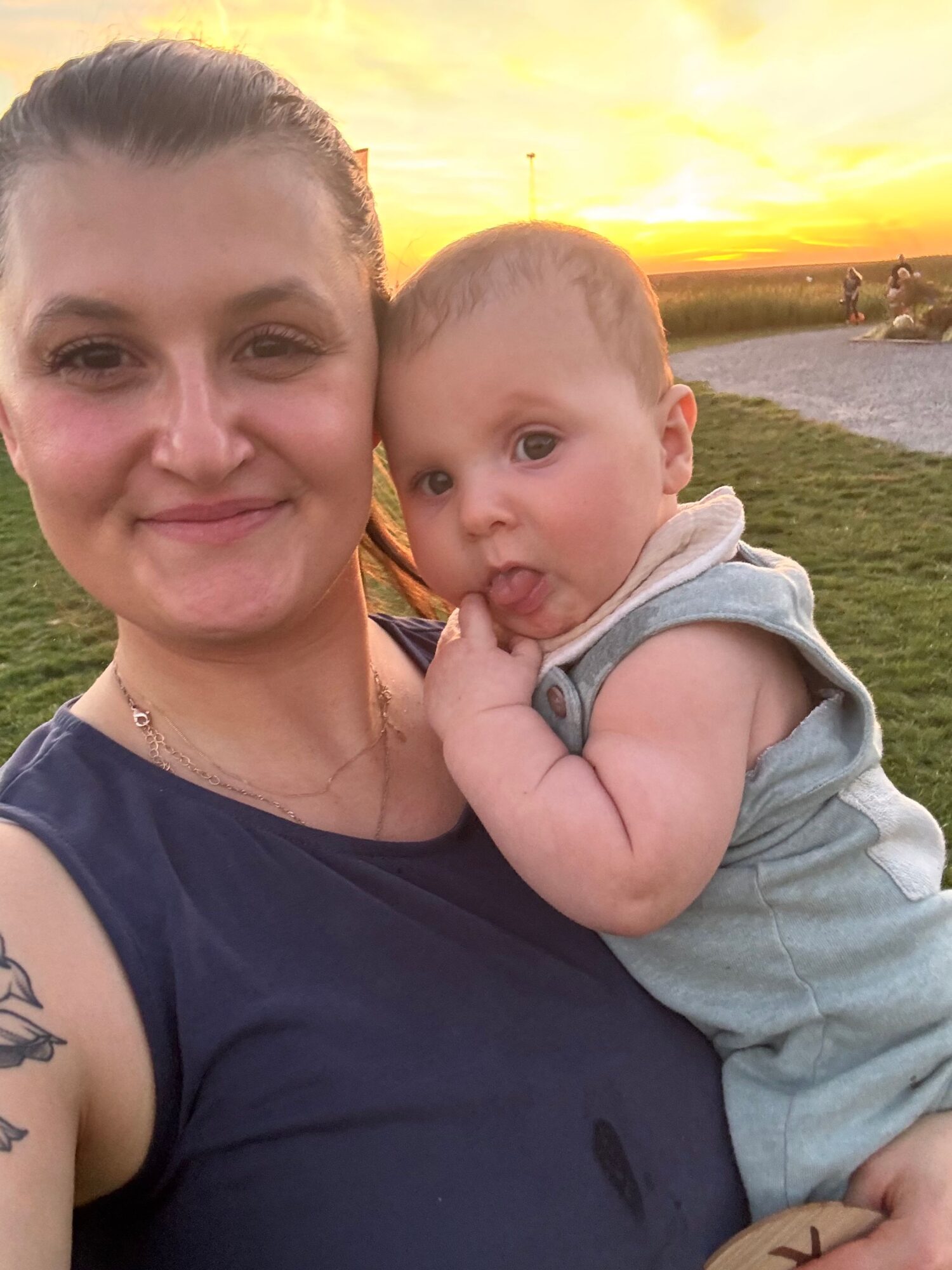 A woman lying on grass with a young child resting on her chest, both smiling, during sunset.
