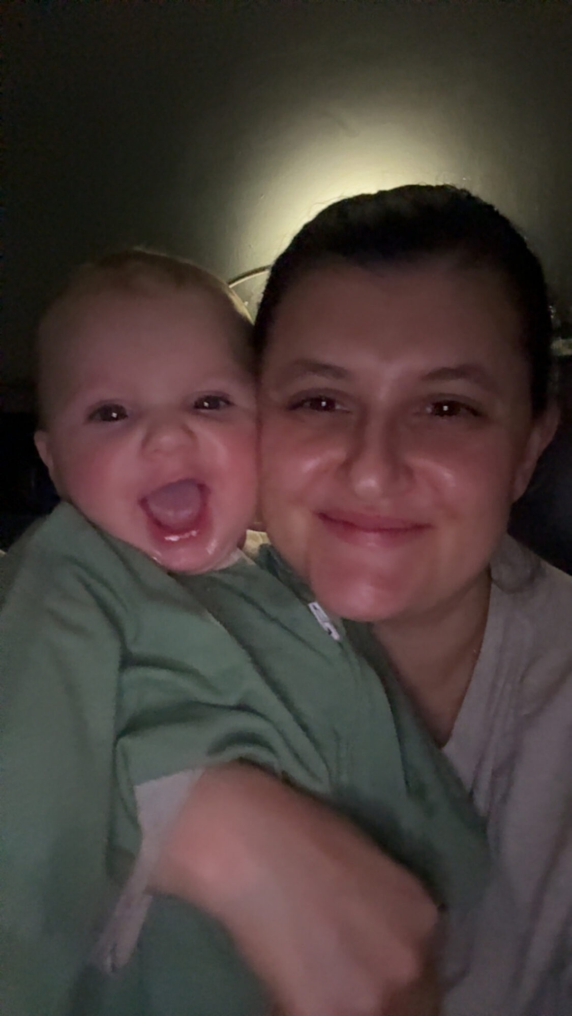 A young person and a baby smiling and close together, with a dark background and light behind them.