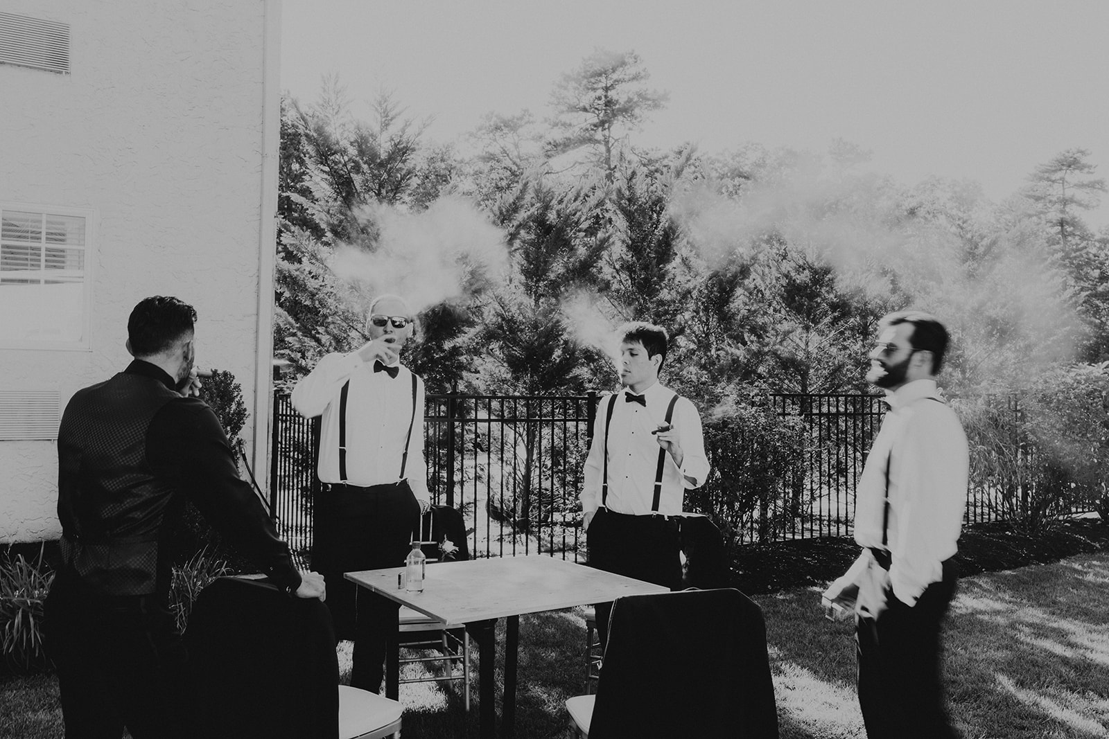 Four men standing outdoors near a table, with trees and a fence in the background, engaged in conversation.