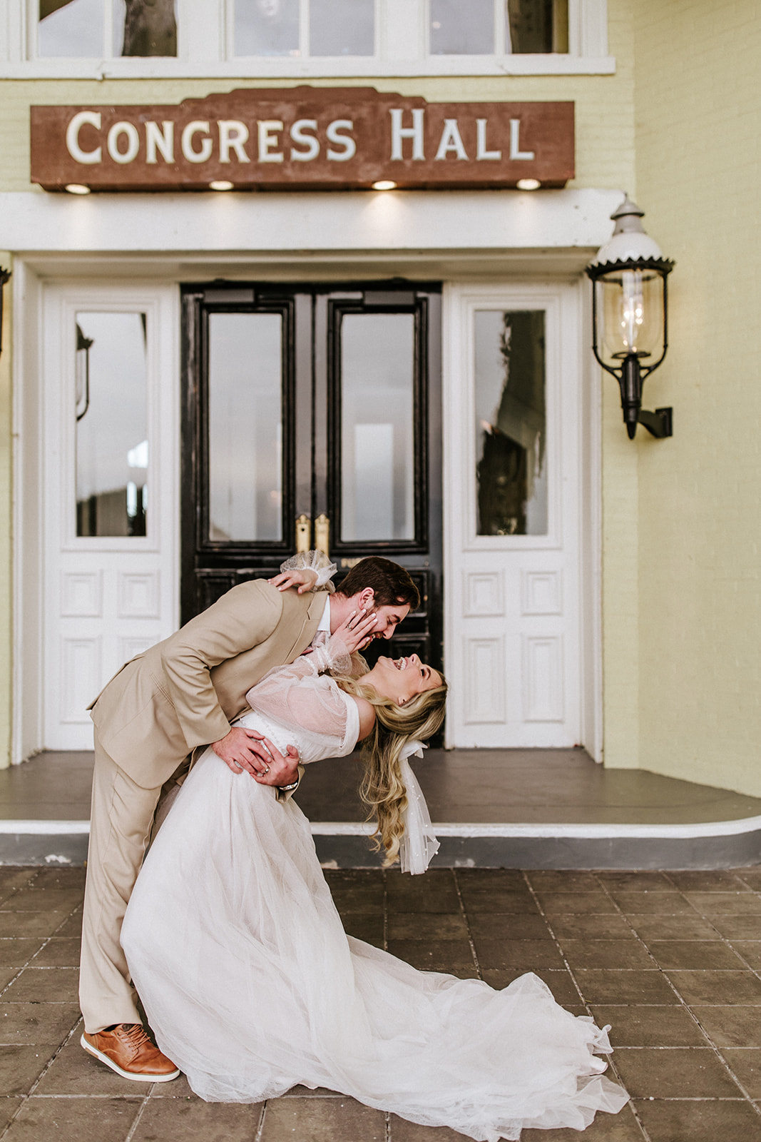 Couple dancing in front of a building with a sign reading 'Congress Hall', with a lantern on the wall, on a tiled porch.