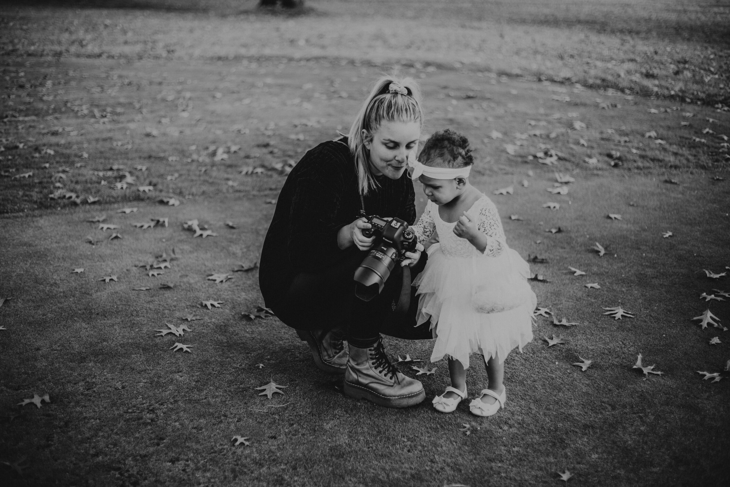 Woman and young girl looking at a camera outdoors with fallen leaves on the ground.