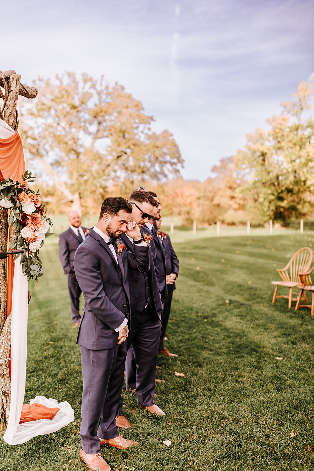 Three men in suits standing outdoors on grass, with trees and chairs in the background, during daytime.
