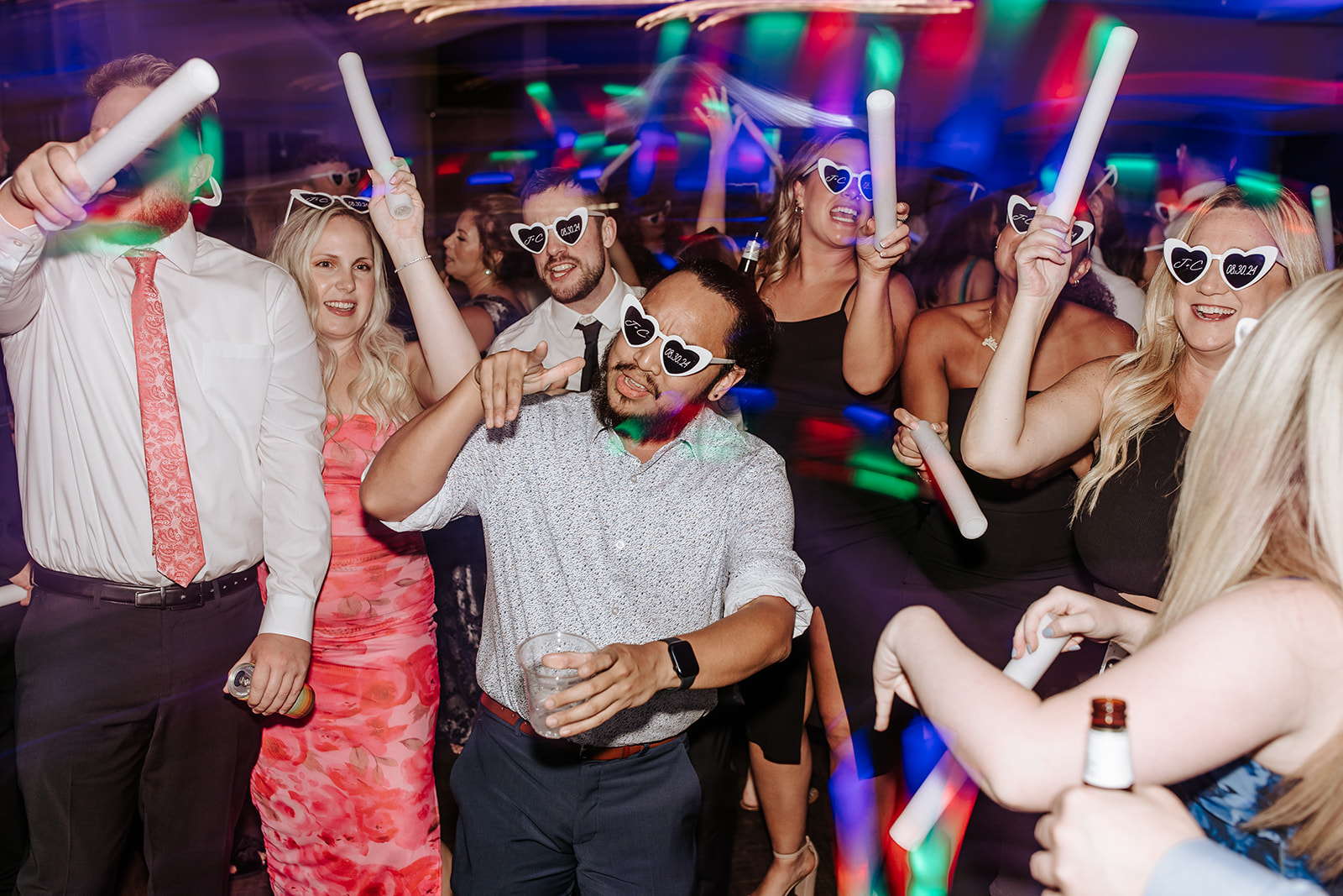 Group of people celebrating with glow sticks, wearing sunglasses, and dancing in a colorful party setting.
