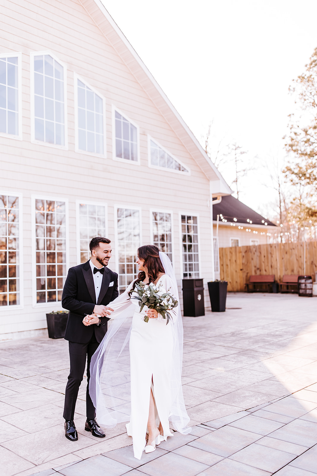 A couple dressed in wedding attire standing outdoors near a large building with multiple windows, holding hands and smiling.
