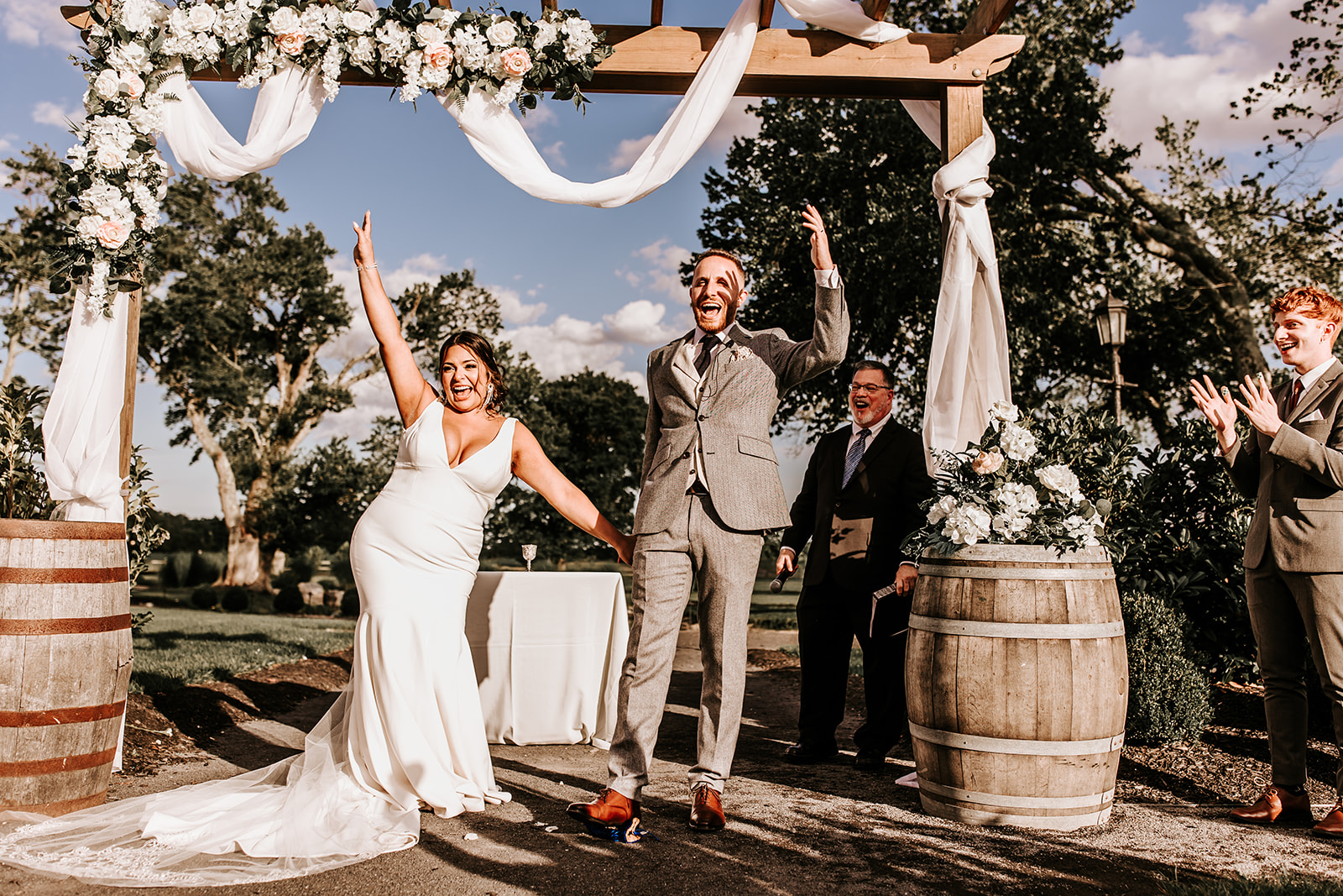 Bride and groom celebrating outdoors under a decorated arch, with guests clapping nearby, trees and sky in background.