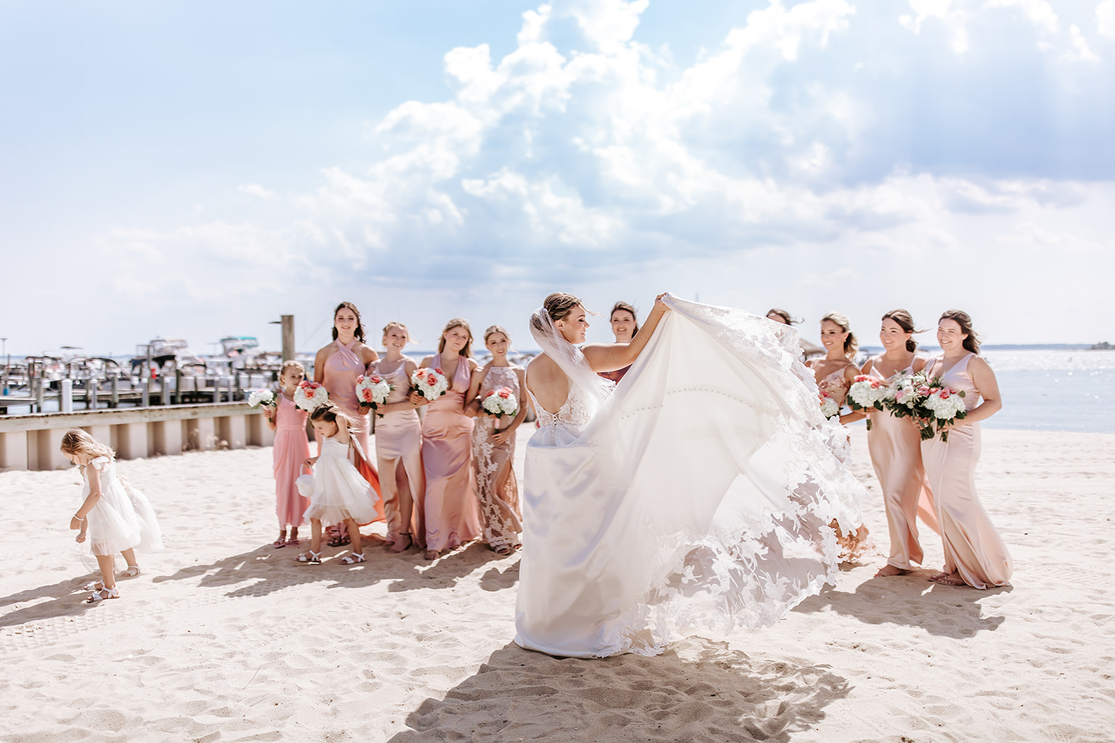Bride in white dress lifts her gown on a sandy beach with bridesmaids in pink dresses holding bouquets, ocean and sky in background.