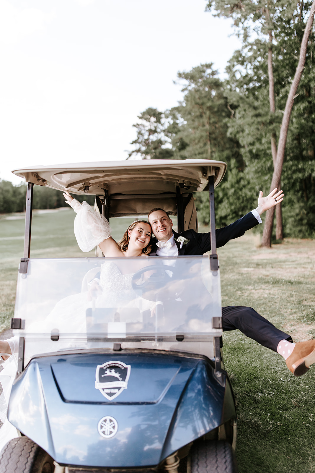 A couple sitting in a golf cart outdoors, smiling, with one person waving and trees in the background.