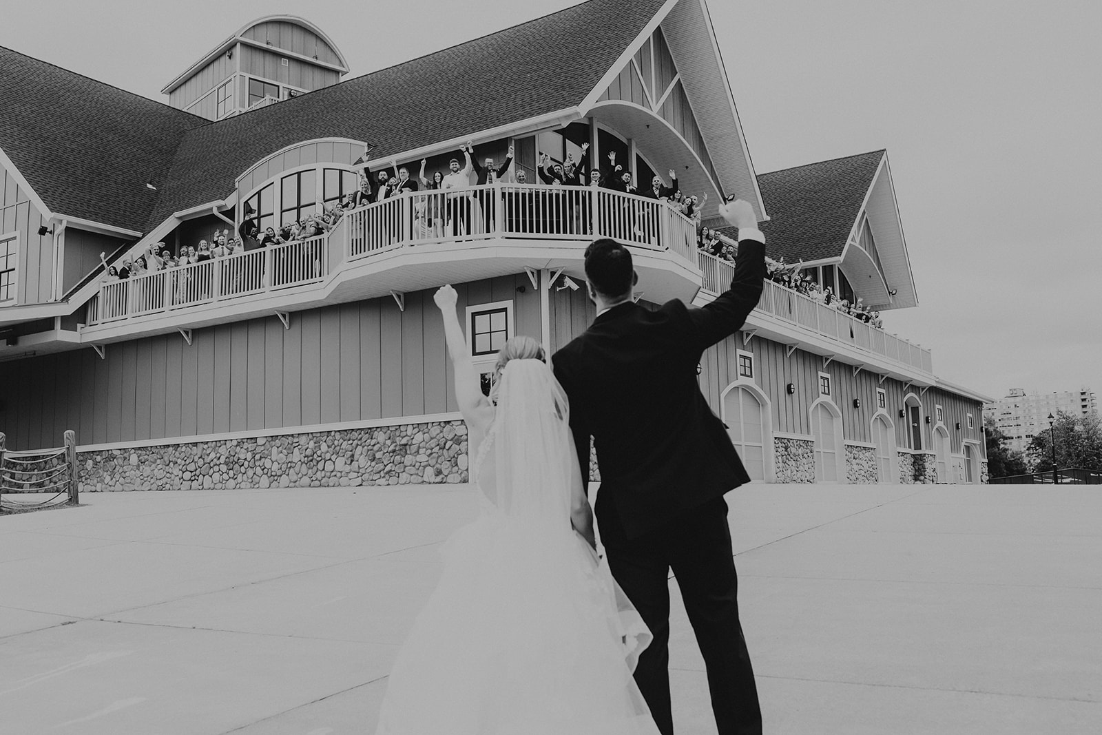 A couple in wedding attire stands outside a large building, with the man raising his arm, in black and white.