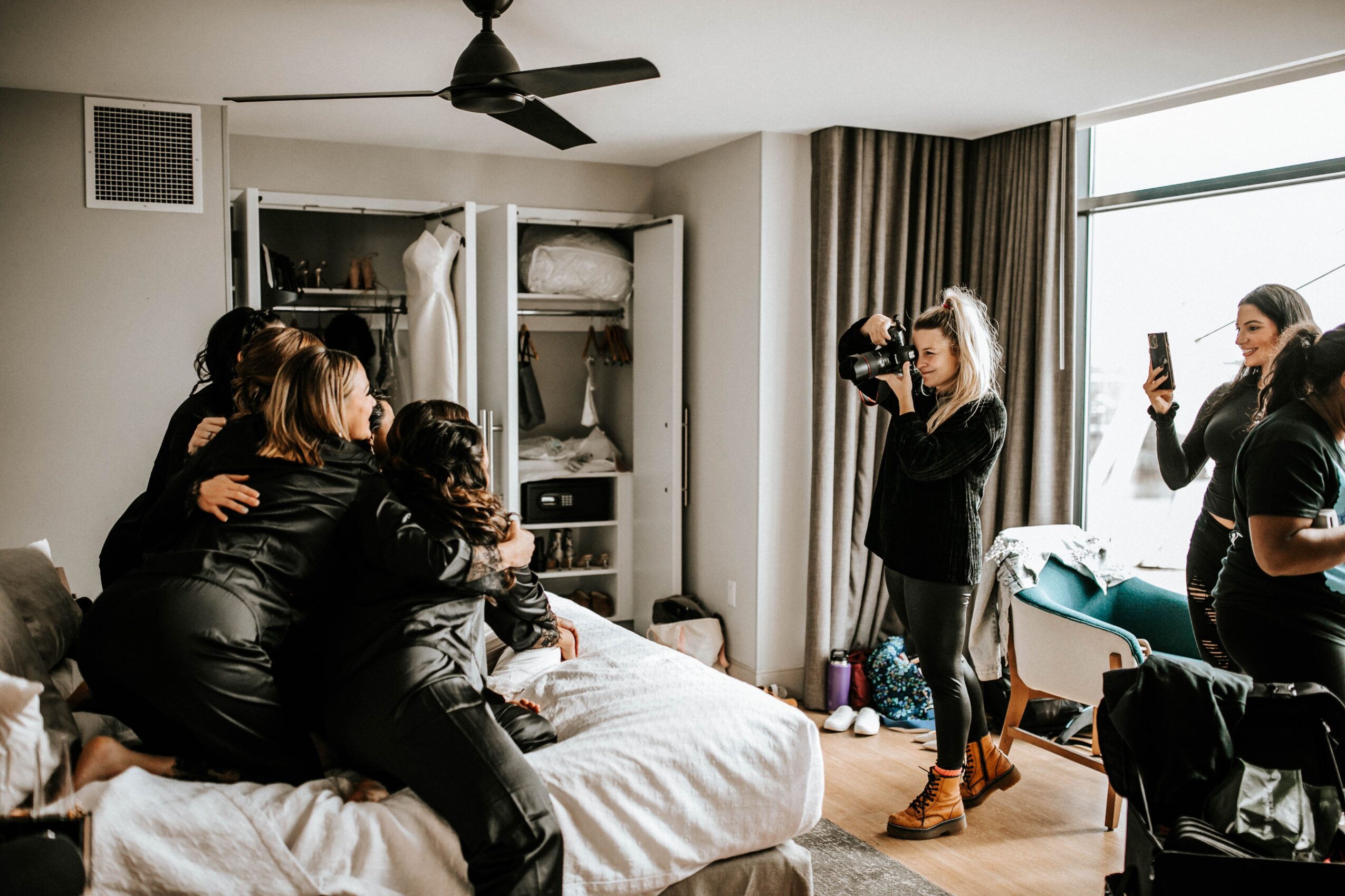 Group of women taking photos and selfies in a bedroom with an open closet, bed, and window with curtains.