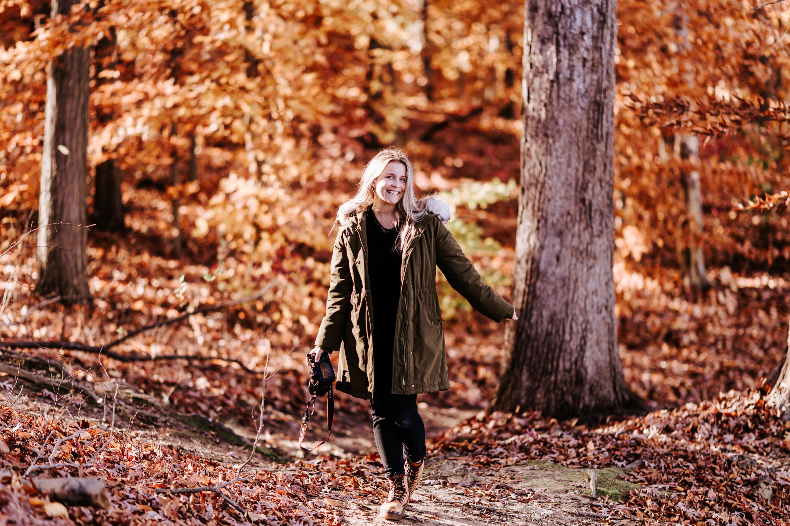 Woman walking in a forest with orange autumn leaves, holding a stick, wearing a coat and glasses.