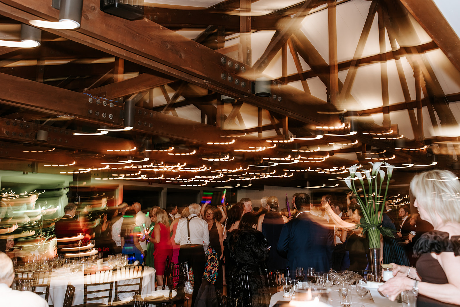 People gather in a large indoor space with wooden beams and string lights, some holding flowers, in a social event or celebration.