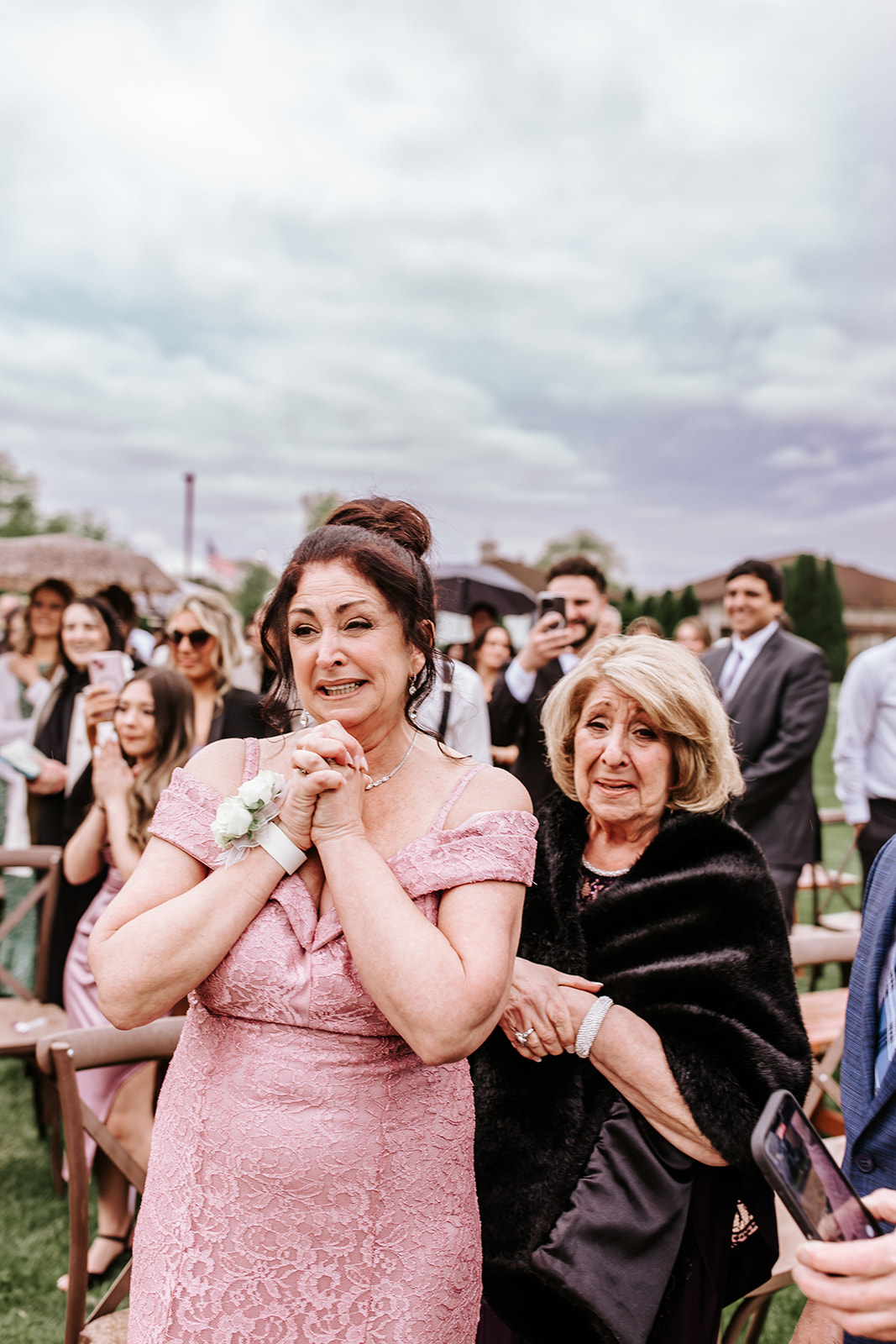 Two women smiling, one in a pink dress and the other in black, at an outdoor event with people in the background.