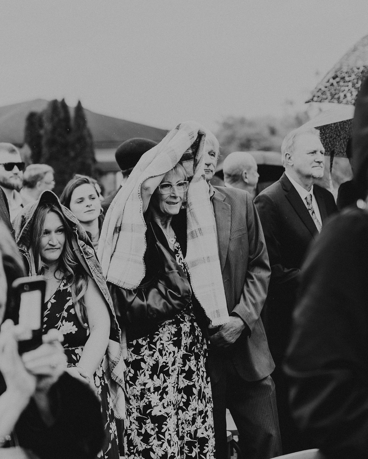 Group of people outdoors, some holding umbrellas, with a woman smiling and holding a hat, in black and white.