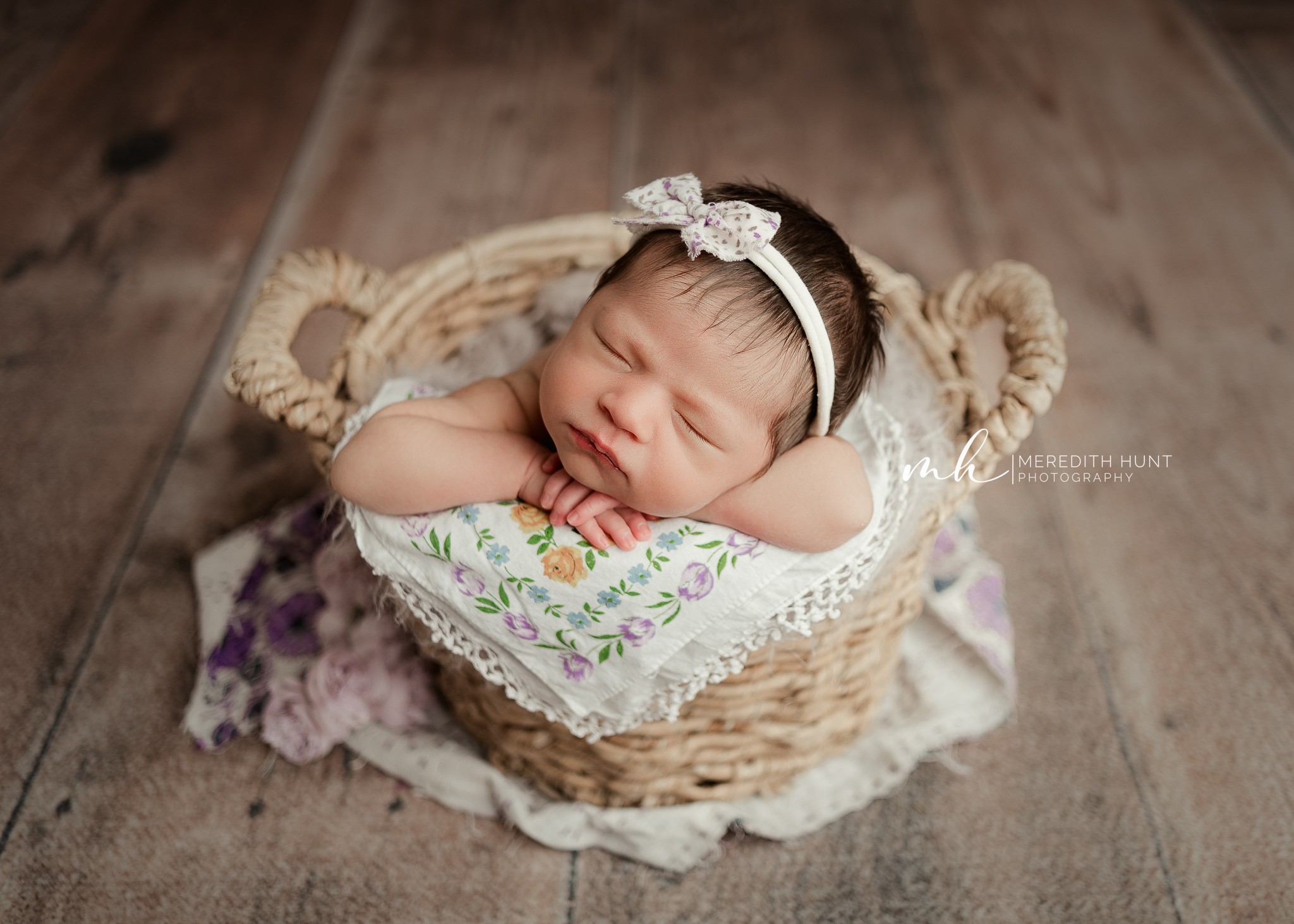 Baby with a headband resting in a woven basket on a wooden floor, surrounded by a cloth with floral pattern.
