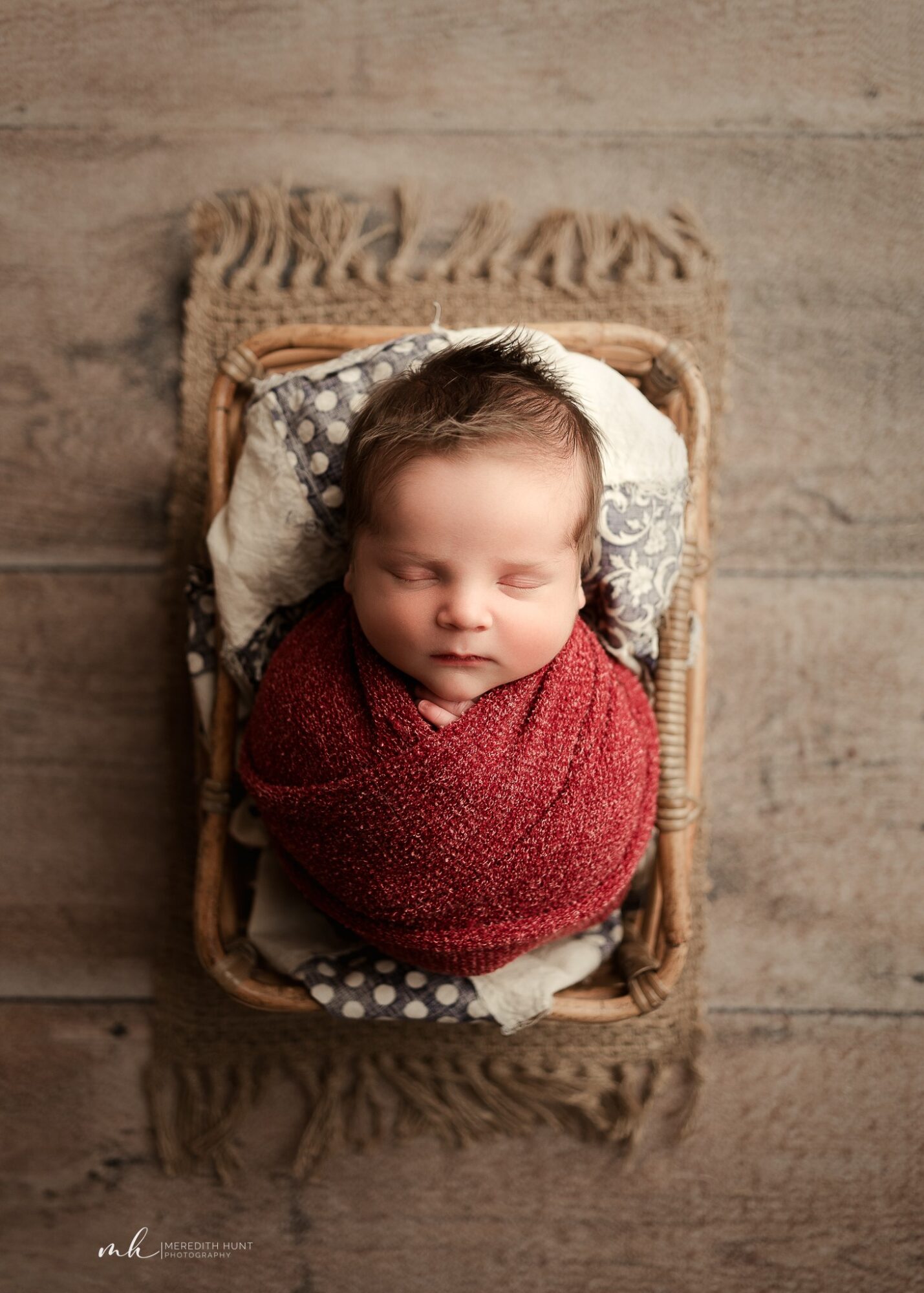 Baby wrapped in a red blanket, lying in a wicker basket on a wooden floor, eyes closed, peaceful expression.