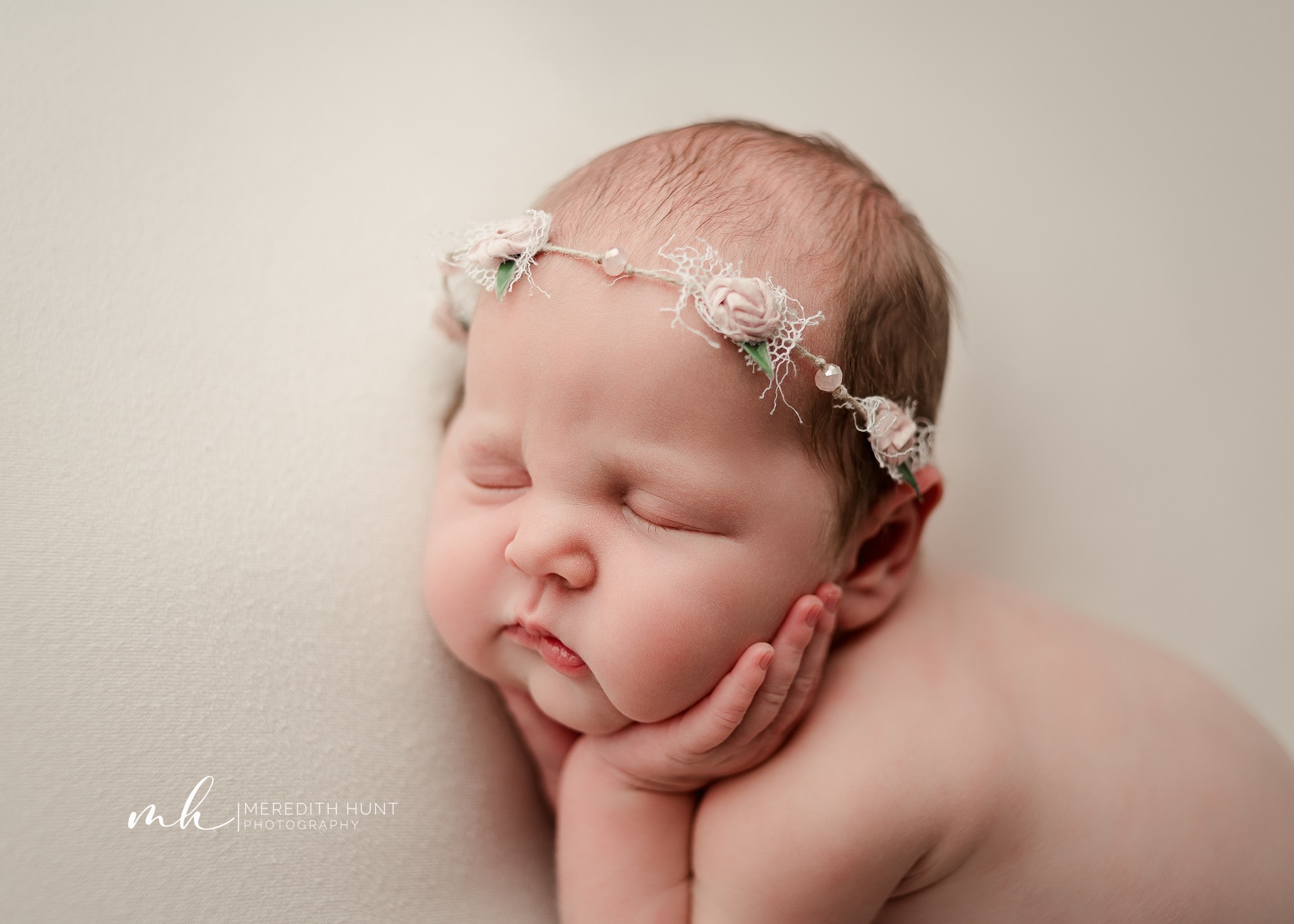 Baby with a floral headband resting face down on hand, eyes closed, against a plain background.