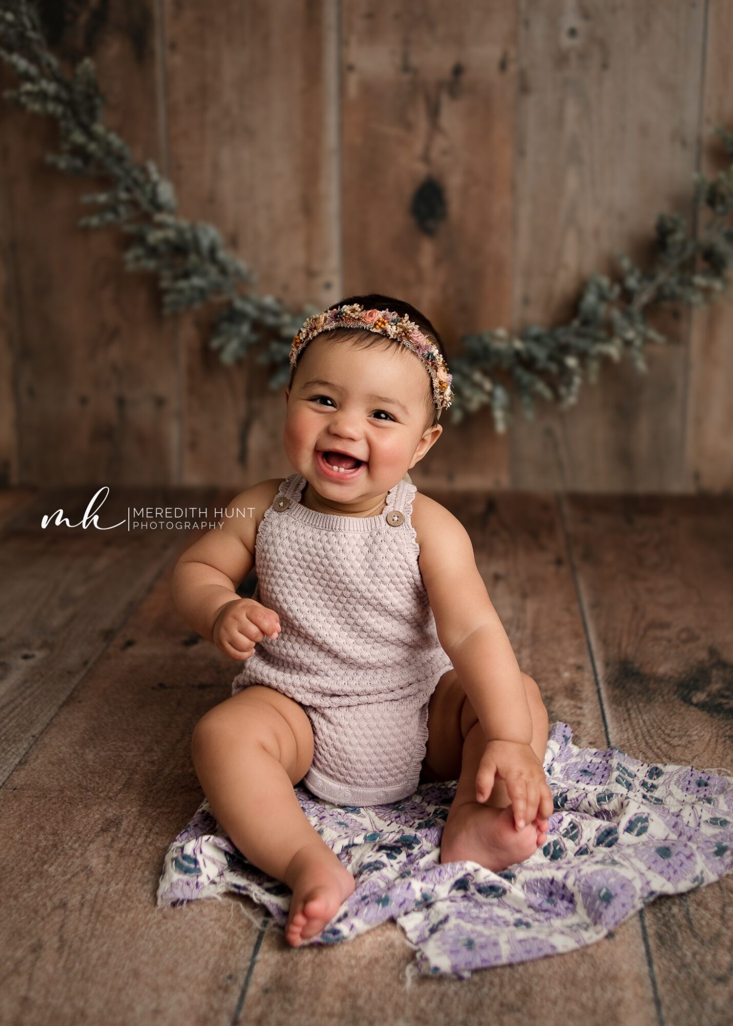 Smiling baby girl sitting on a blanket with a wooden background and greenery, wearing a polka dot outfit and headband.