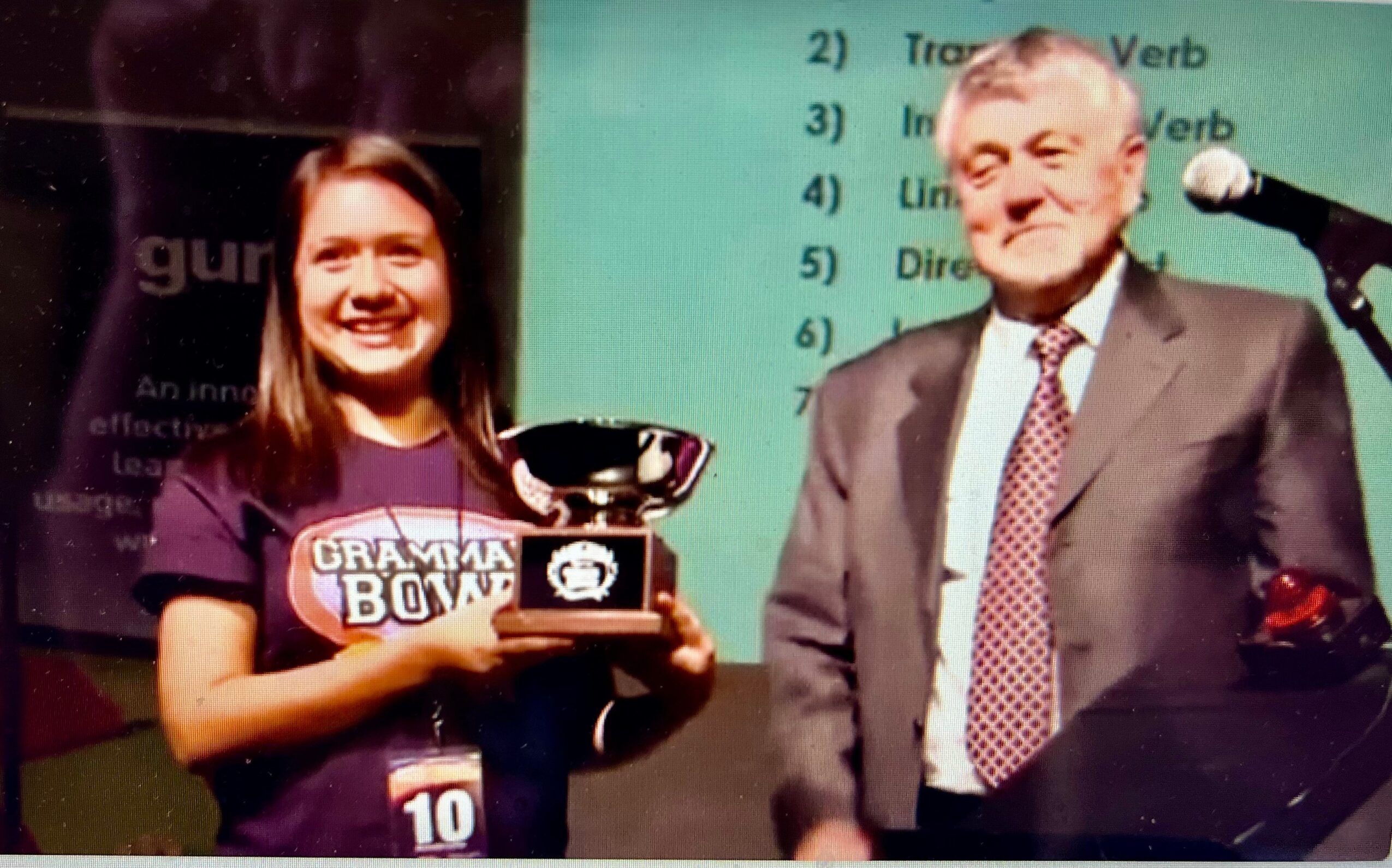 Young woman holding a trophy on stage with an older man in a suit, background shows a presentation slide.