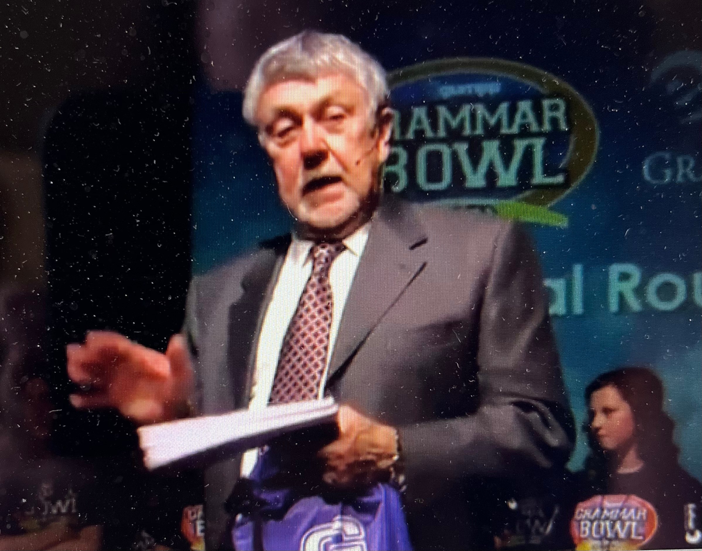 Older man in suit speaking at event, holding papers, with a woman in background, and a sign reading 'GAMMAR BOWL'.