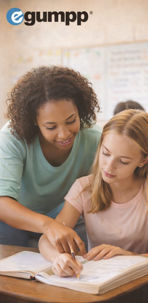 Two women look at a book together in a classroom, one pointing at the pages, with a whiteboard in the background.