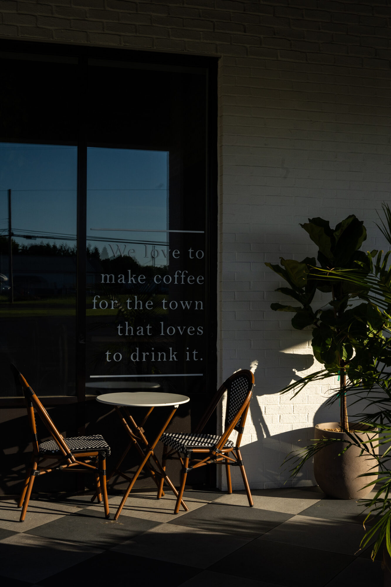 Outdoor seating with two chairs and a small table near a window with text, potted plant nearby, sunlight casting shadows.