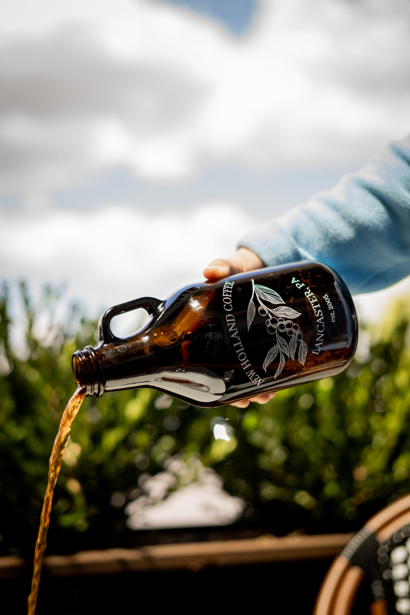 Person pouring dark beer from a bottle outdoors with a cloudy sky and greenery in background.