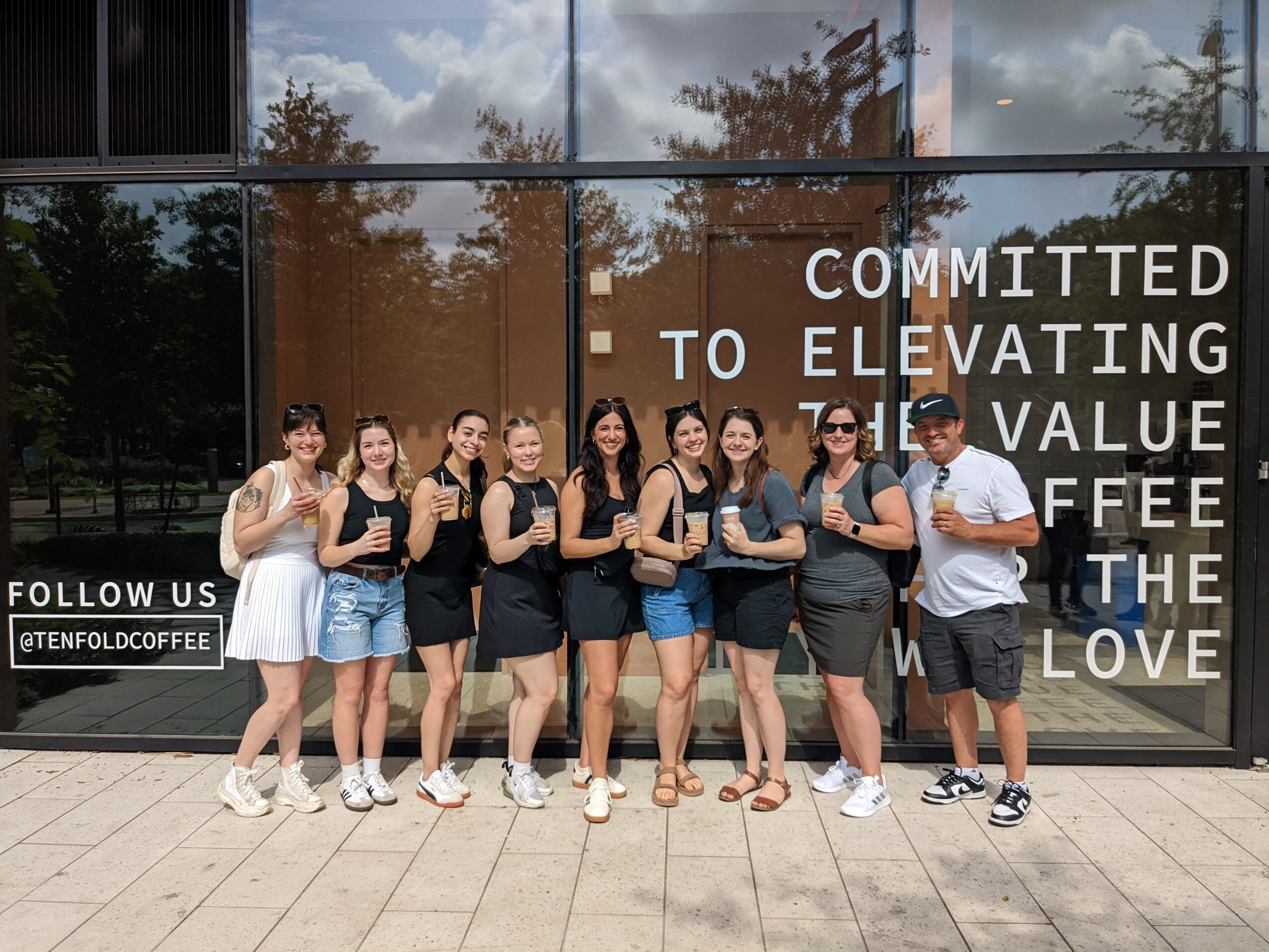 Group of nine people standing outside in front of a glass window with text, smiling and posing for the photo.