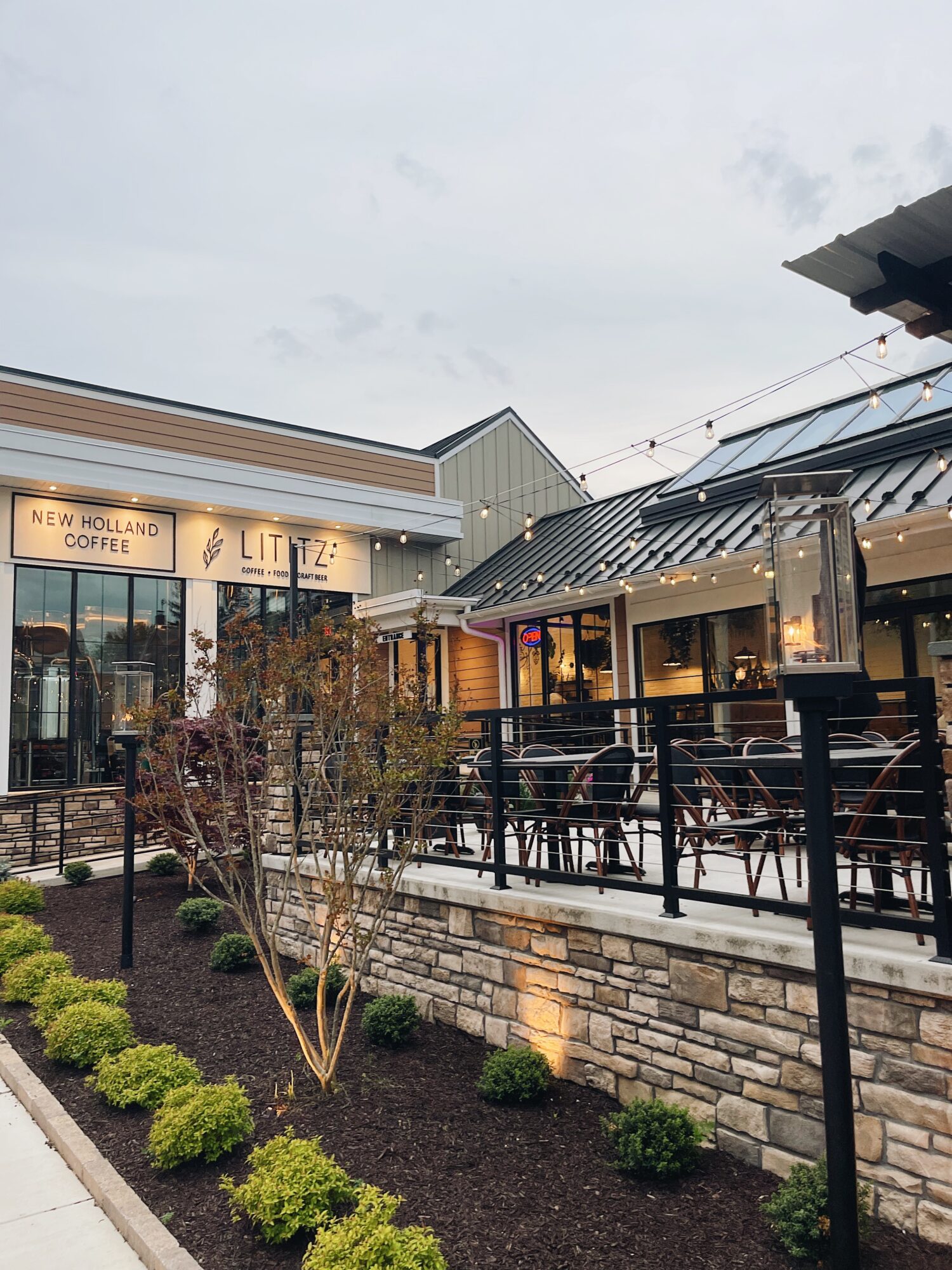 Outdoor seating area with tables and chairs, a building with a sign, and string lights, under a cloudy sky.