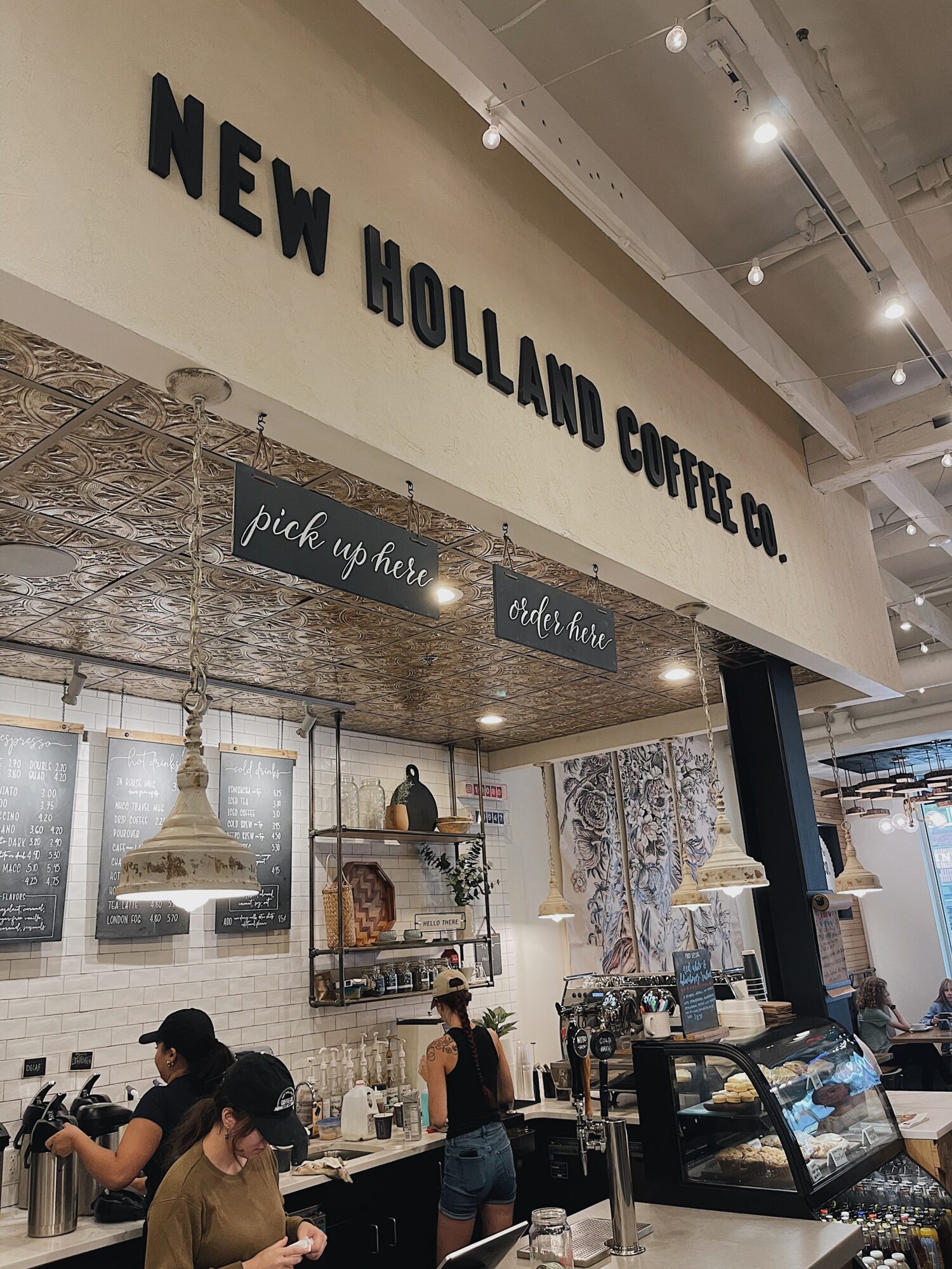 Interior of a coffee shop with people ordering and working, shelves with items, and a display case, under a sign reading 'New Holland Coffee Co.'