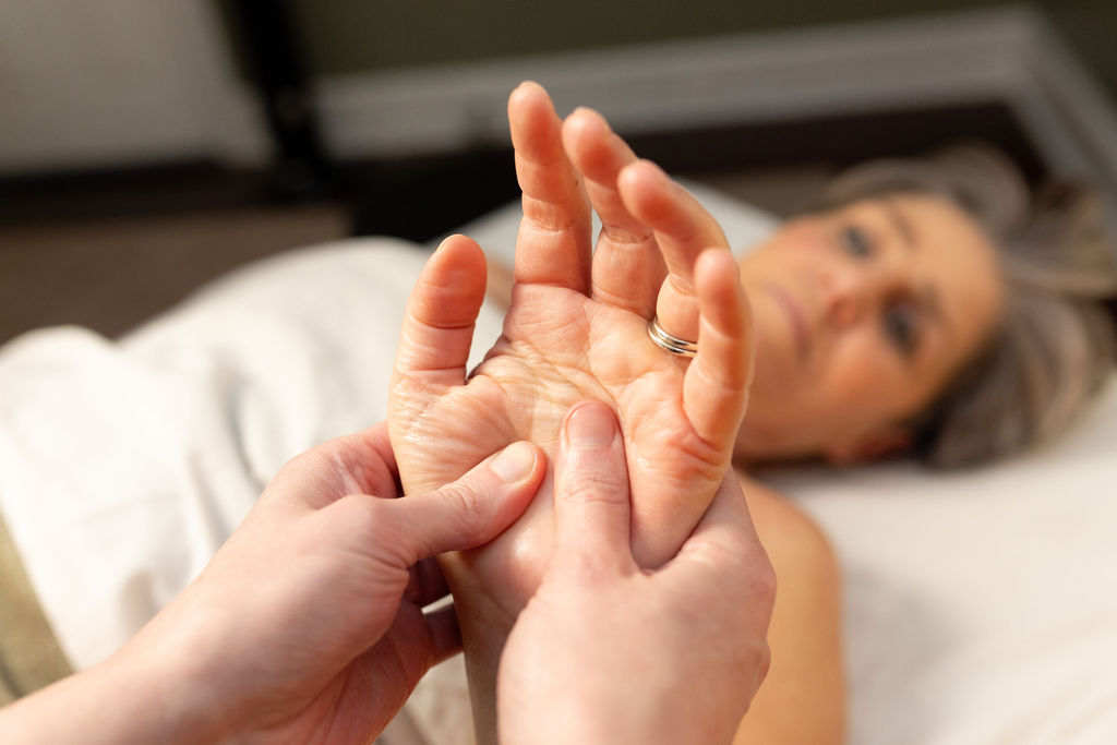 Person lying on bed with hands in foreground, one hand holding the other's fingers, blurred face in background.