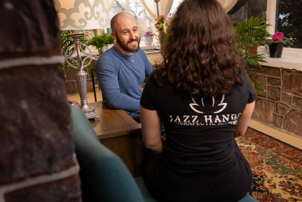 Man with a beard smiling at a woman with curly hair sitting at a table in a cozy room with plants and a patterned rug.