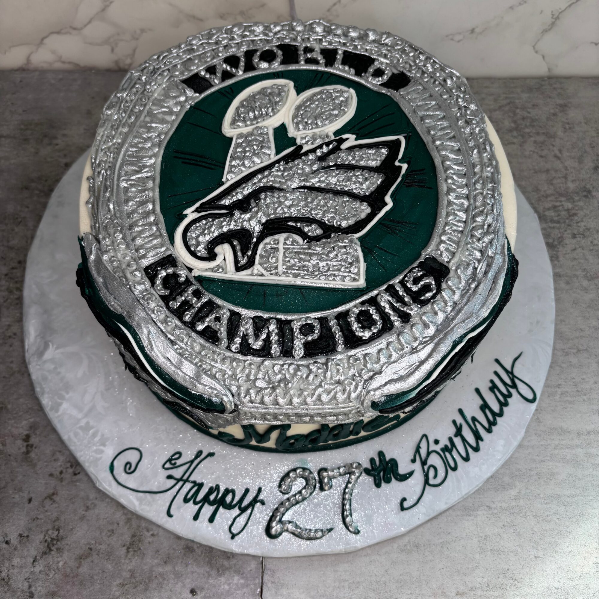 Round cake with Eagles logo, silver and green decorations, and a message reading 'Happy 27th Birthday' on a white base.