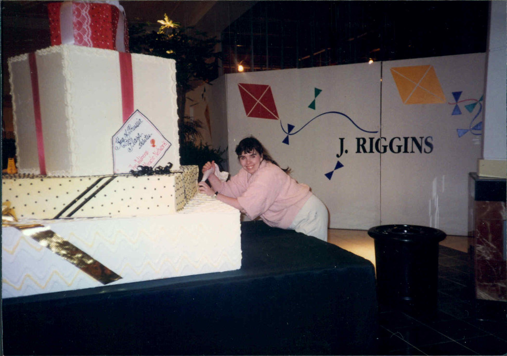 Person leaning on a table with large gift boxes, a J. Riggins sign, and a black trash can nearby.