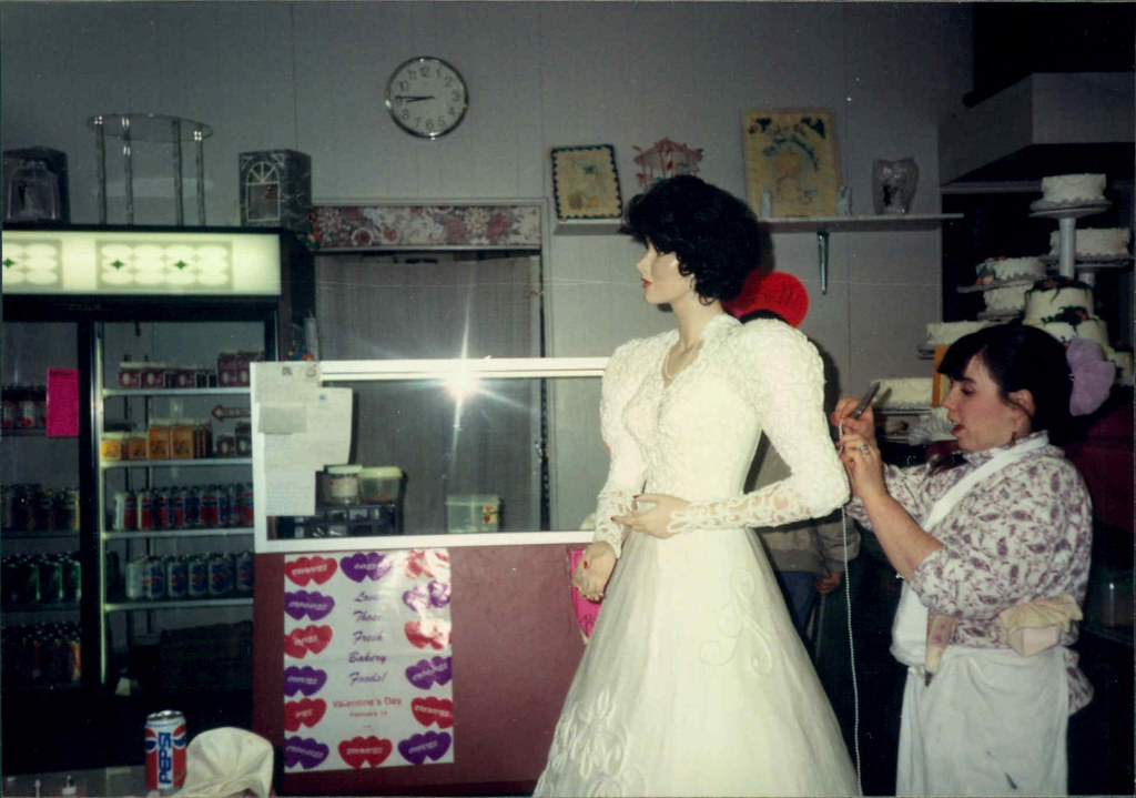 Two women in a store, one in a white dress and the other adjusting her hair, shelves and a clock in background.