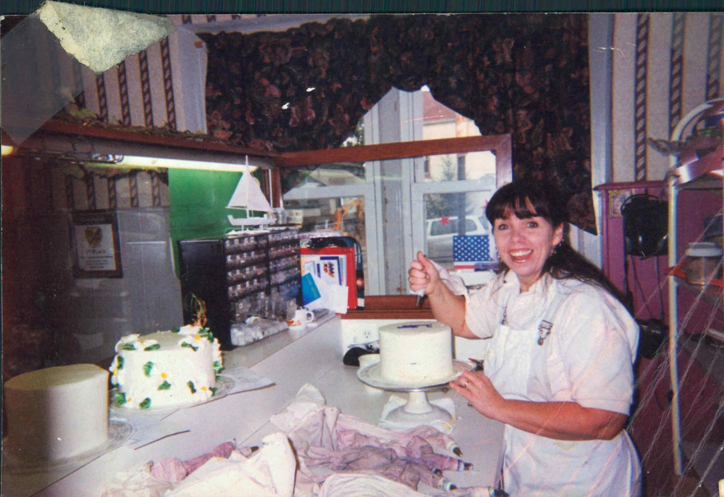 Woman smiling at a cake on a counter, with birthday decorations and a window in the background.