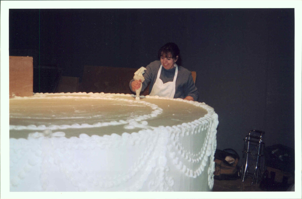 Person decorating a large, round cake with white frosting, standing behind it, in a dark room.