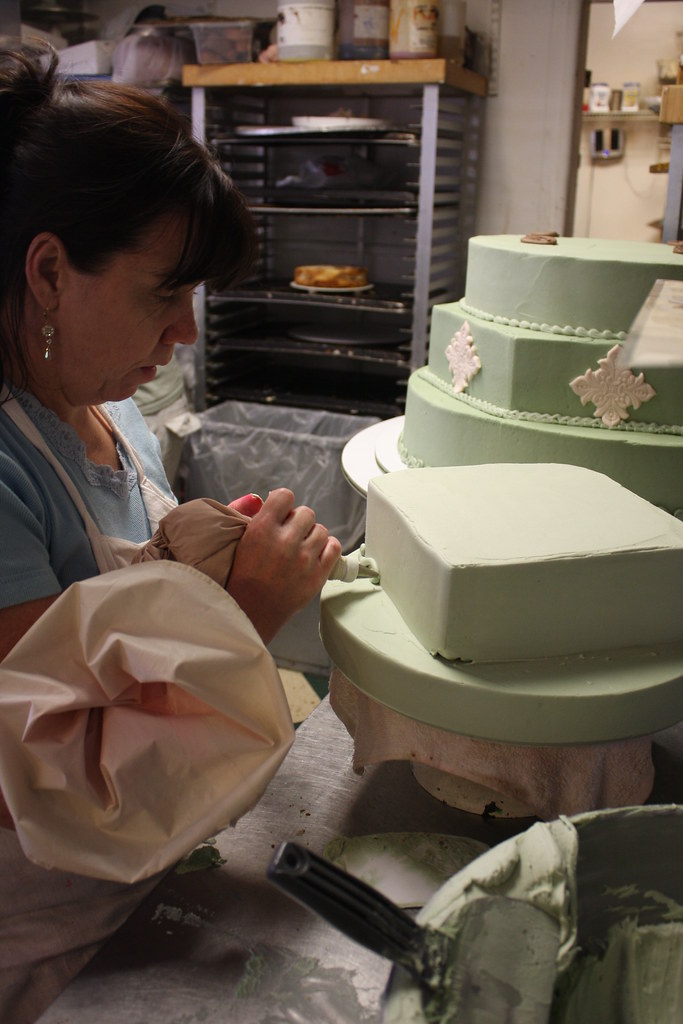 Woman decorating a tiered cake with green fondant, in a kitchen setting, with baking tools and ingredients visible.